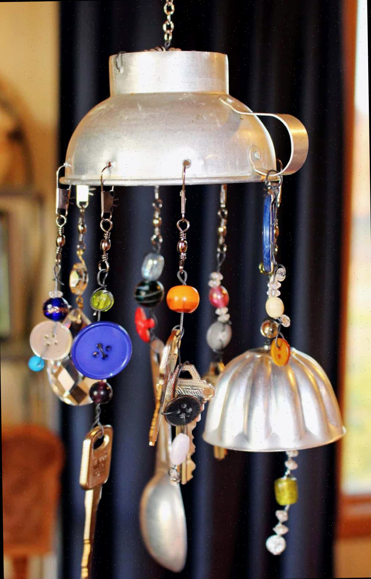 Um sino de vento DIY finalizado confeccionado com contas e objetos de cozinha vintage pendurado em um espaço interno.