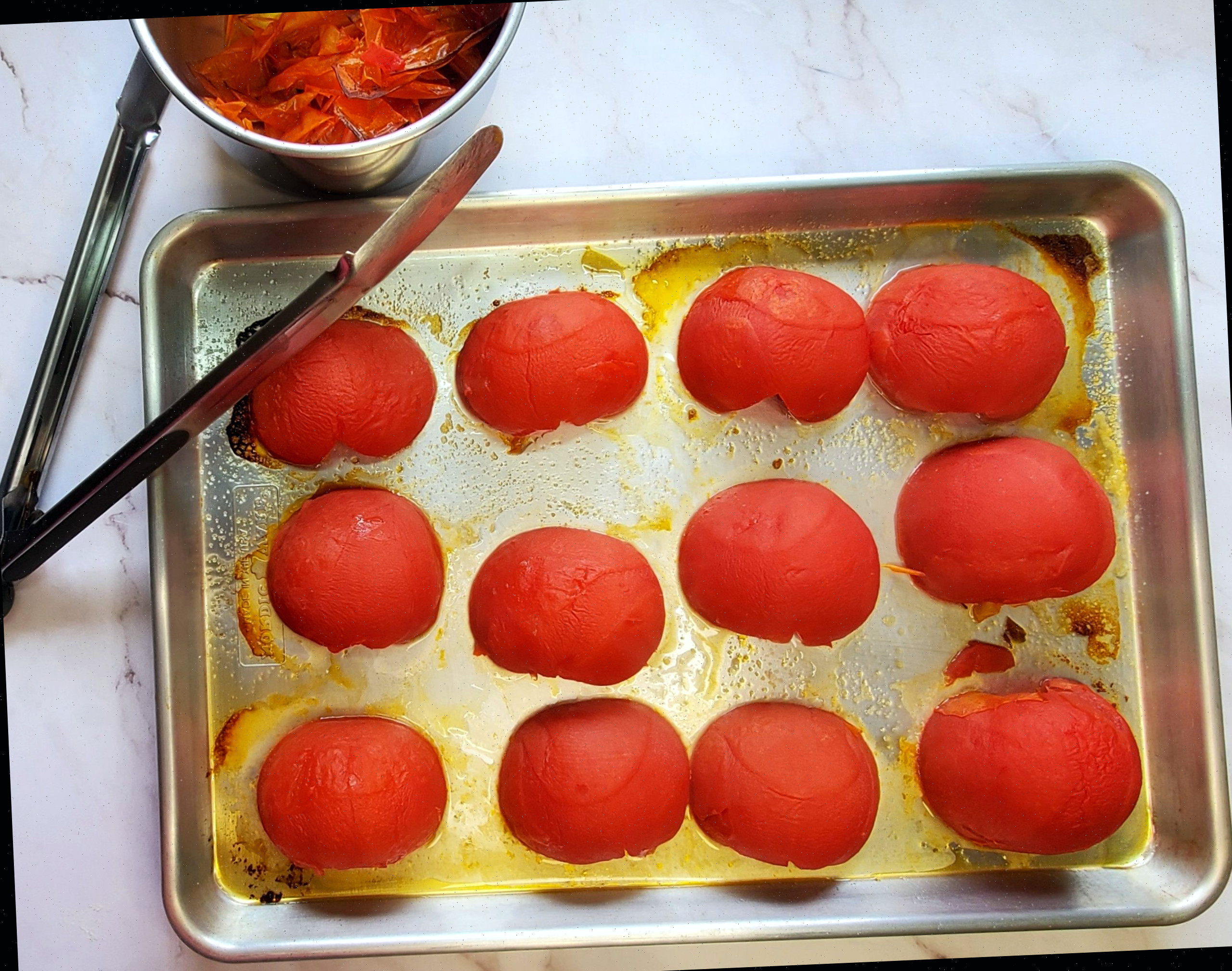 Tomates beefsteak assados com uma pinça descansando na assadeira, ao lado de uma tigela com cascas de tomate.