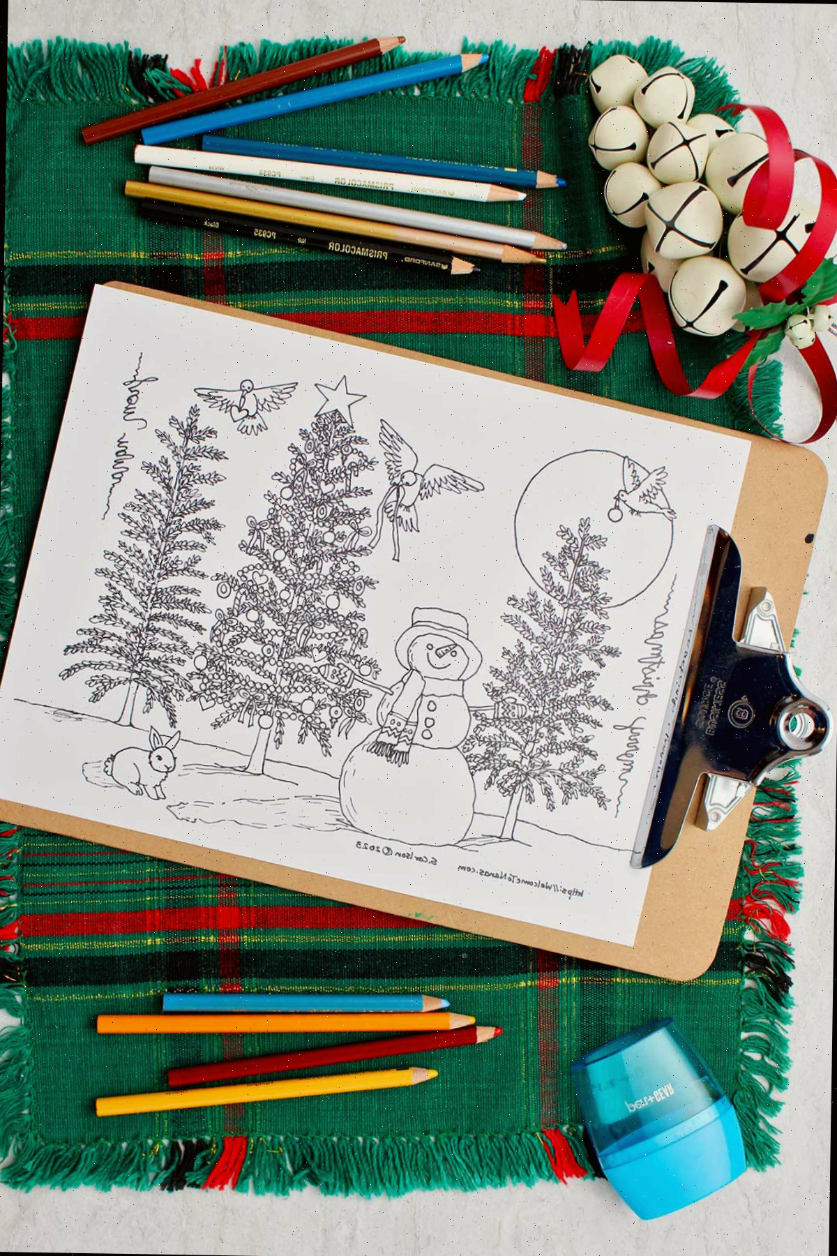 Página para colorir árvore de natal sem coloração em um clipboard repousando sobre um local de mesa verde e vermelho com lápis de cor, um apontador e sinos ao lado.