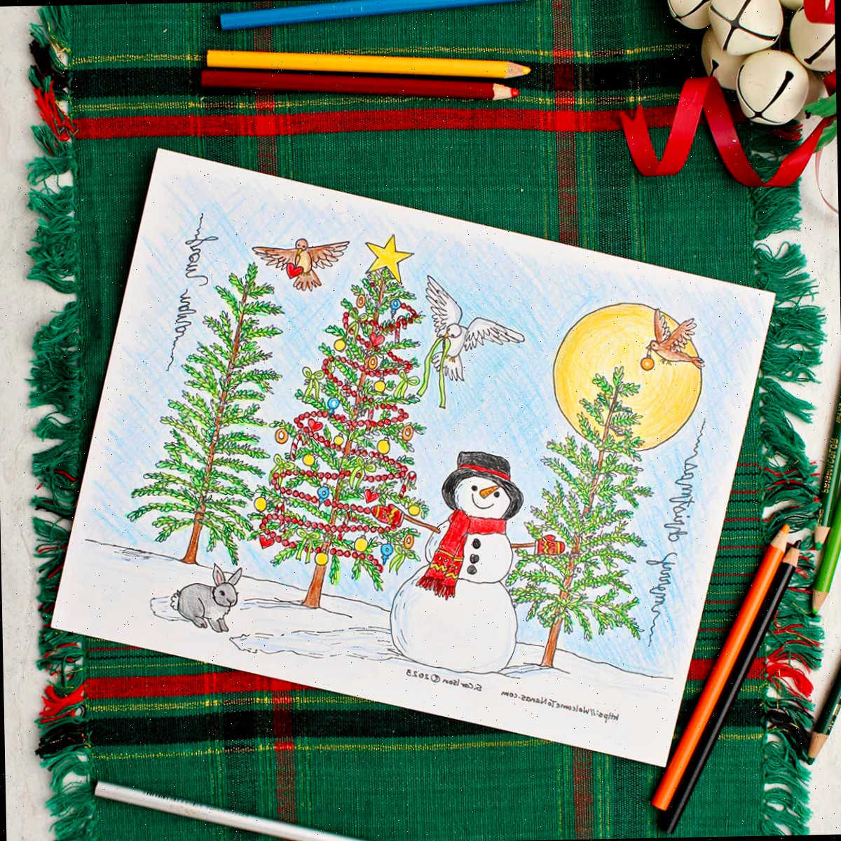 Página de colorir de árvore de Natal completada descansando sobre um caminho de mesa verde e vermelho com sinos decorativos brancos por perto. Imagem quadrada.