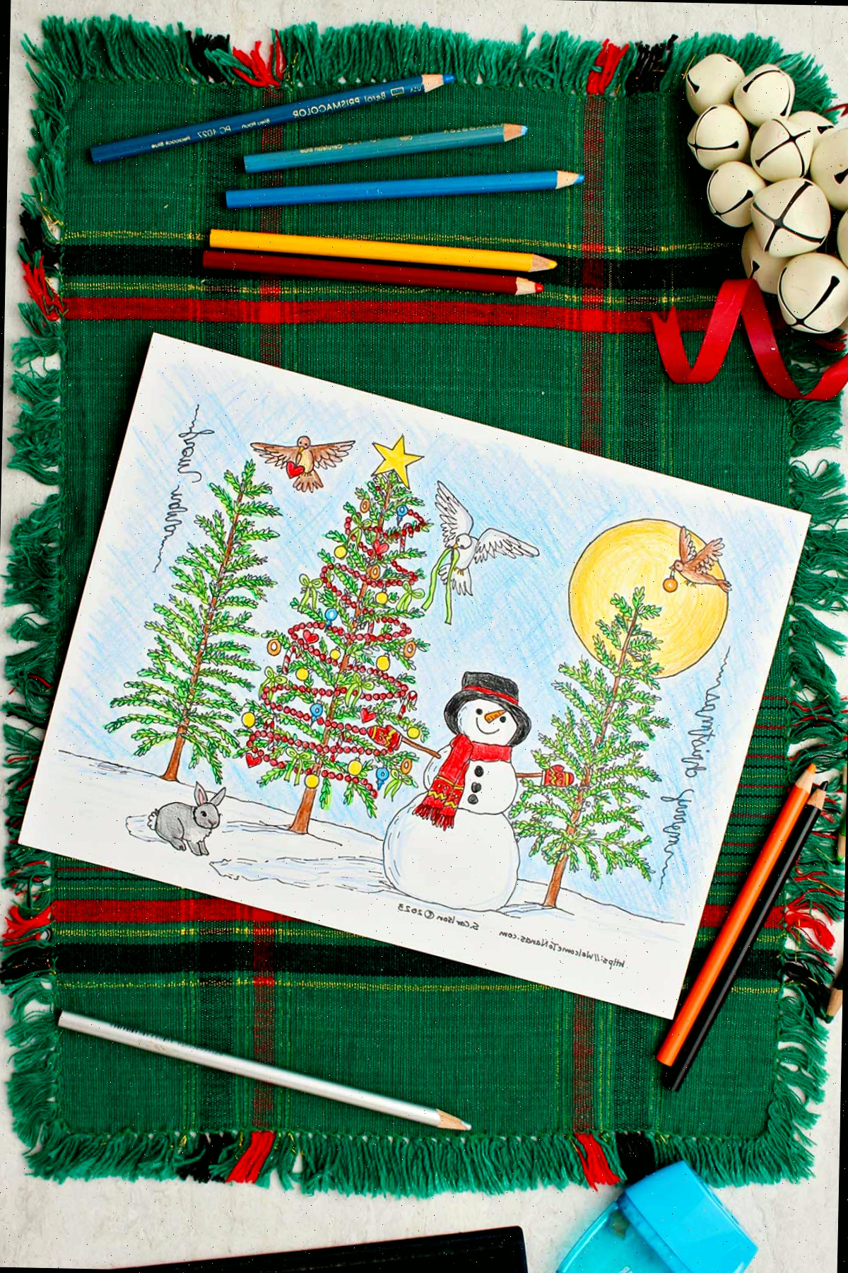 Página para colorir árvore de natal completa repousando sobre um local de mesa verde e vermelho com sinos decorativos brancos e lápis de cor ao lado.