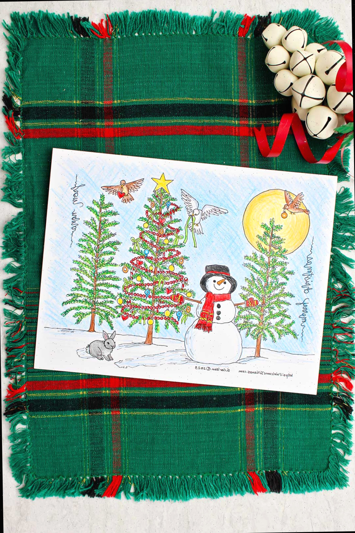 Página de colorir da árvore de natal completa repousando sobre uma toalha de mesa verde e vermelha com sinos decorativos brancos ao redor.