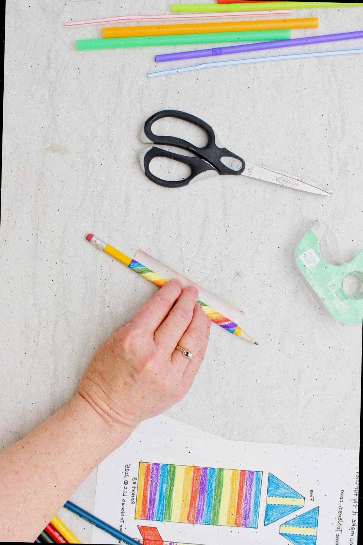 Indivíduo enrolando um molde retangular de papel colorido arco-íris em torno de um lápis.