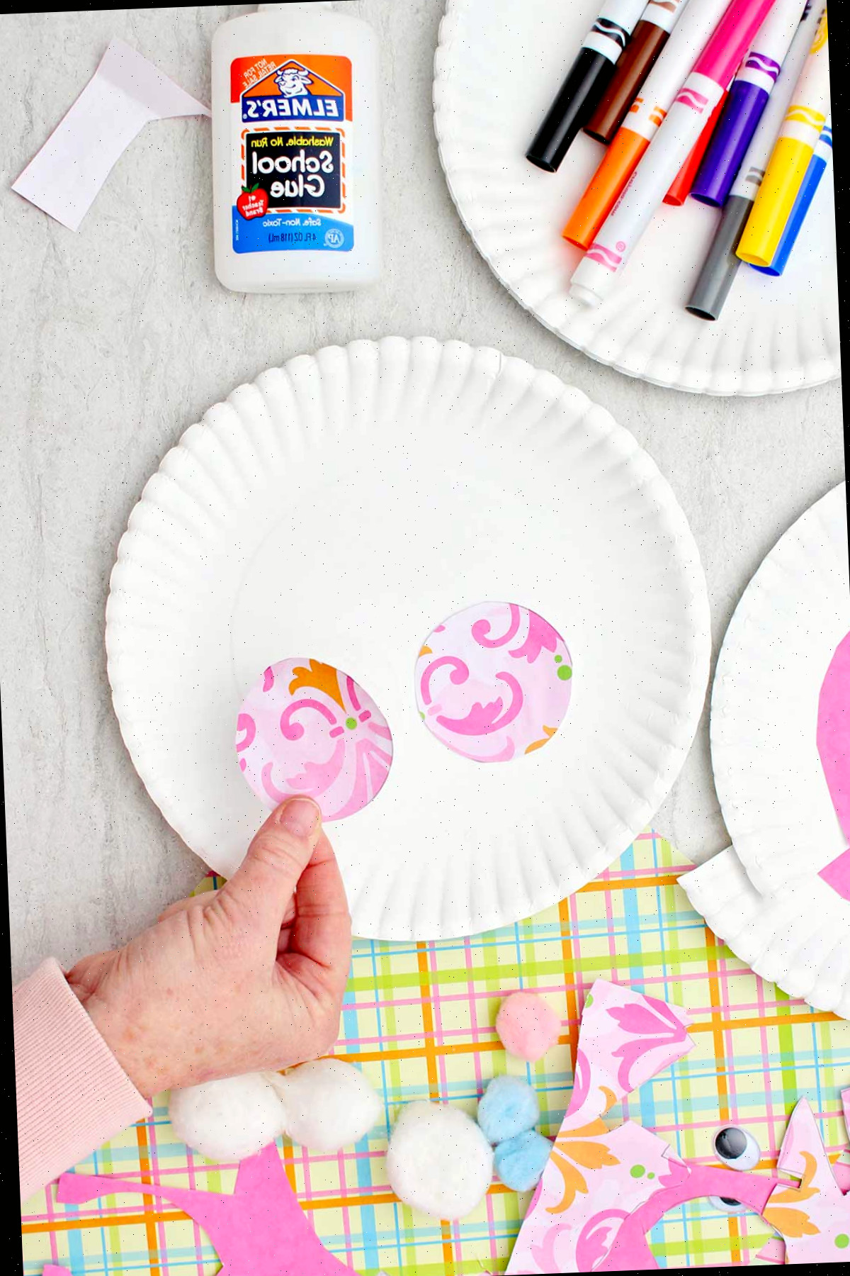 Indivíduo colando dois círculos rosa estampados na parte inferior de um prato de papel para criar as bochechas do coelho, com outros suprimentos próximos.