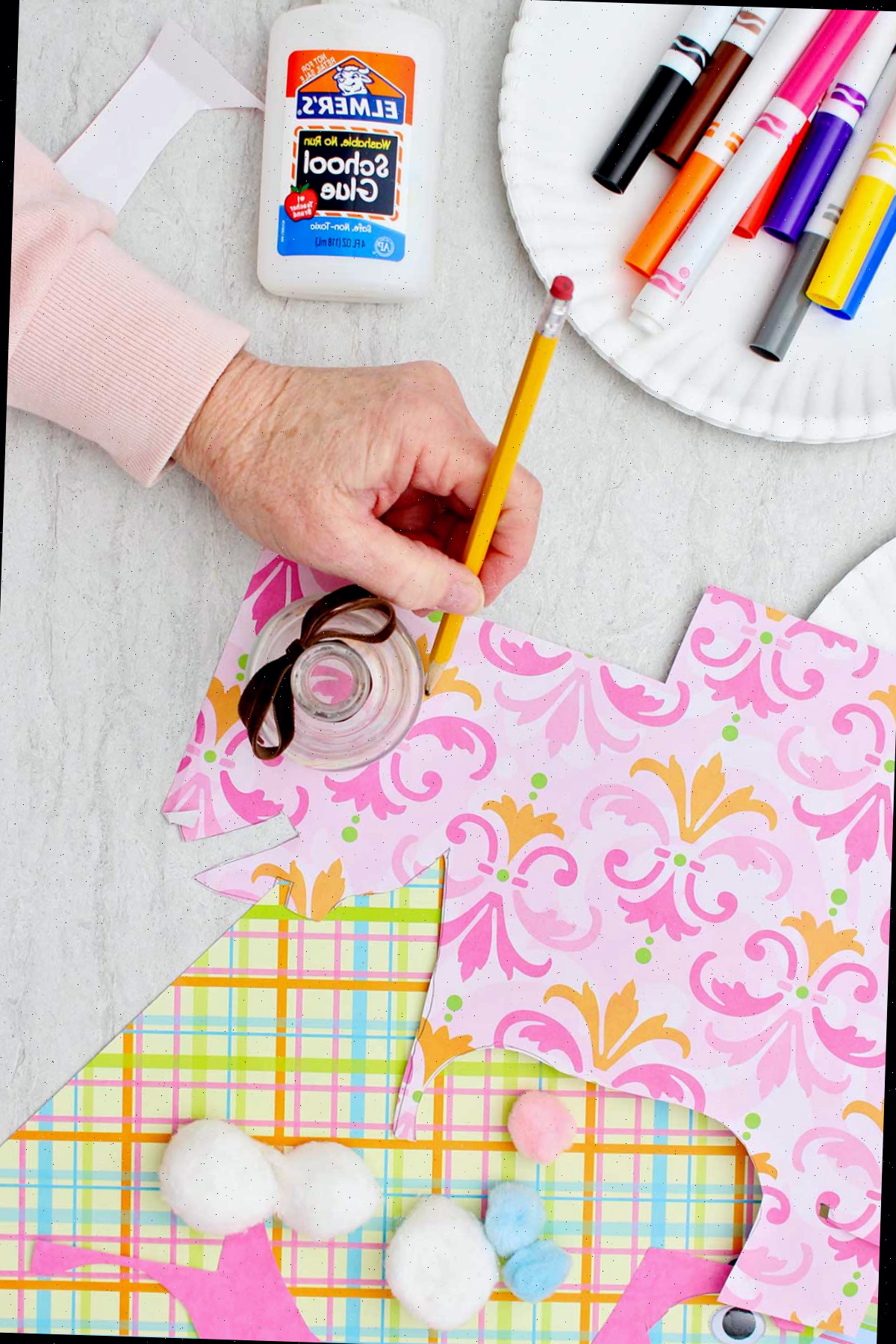 Indivíduo utilizando uma garrafa de vidro para traçar um círculo em um pedaço de papel rosa estampado, com outros materiais nas proximidades.