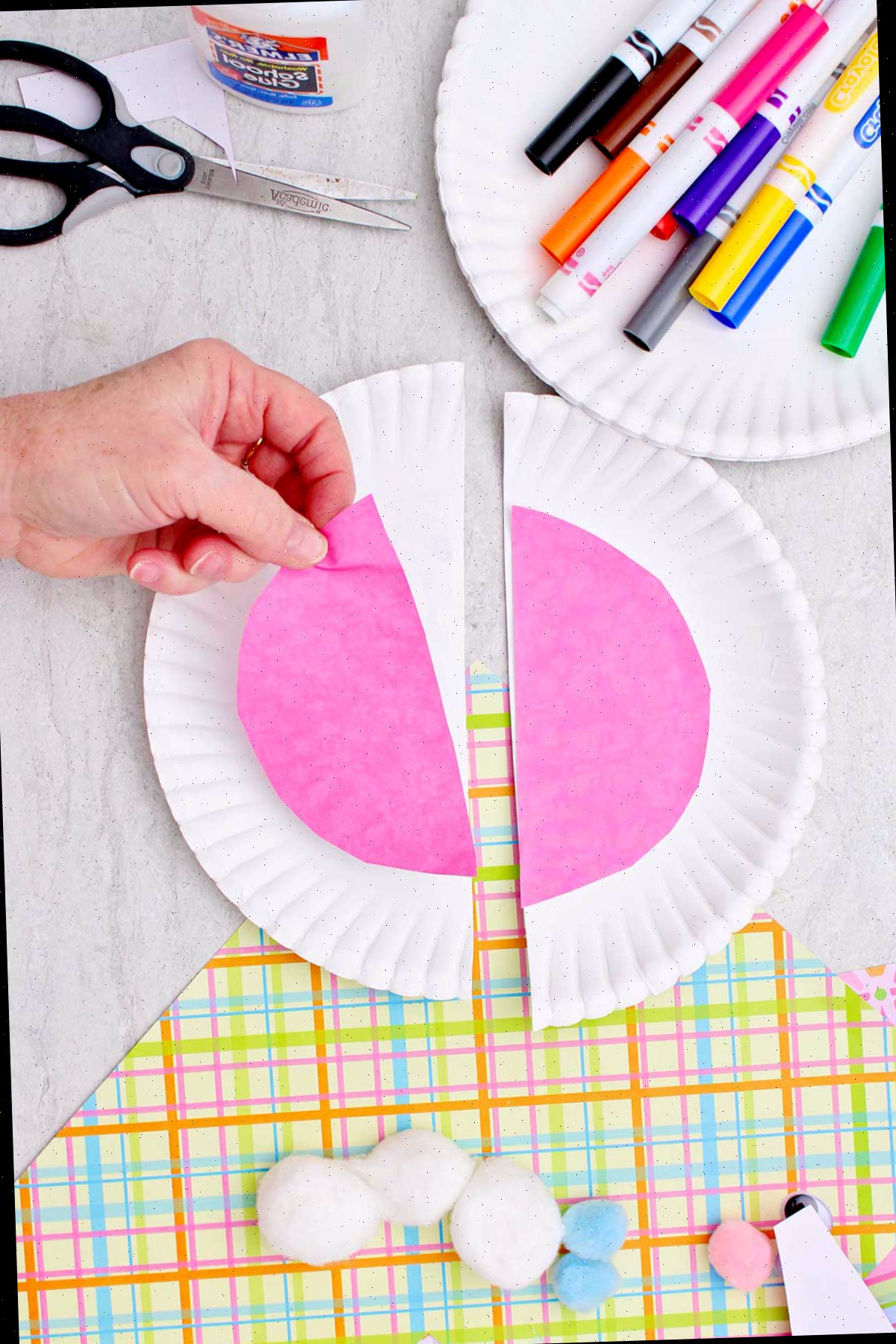 Pessoa fazendo um corte em forma de meio círculo em um pedaço de papel rosa sobre uma metade de um grande prato, para criar orelhas de coelho, com outros materiais nas proximidades.