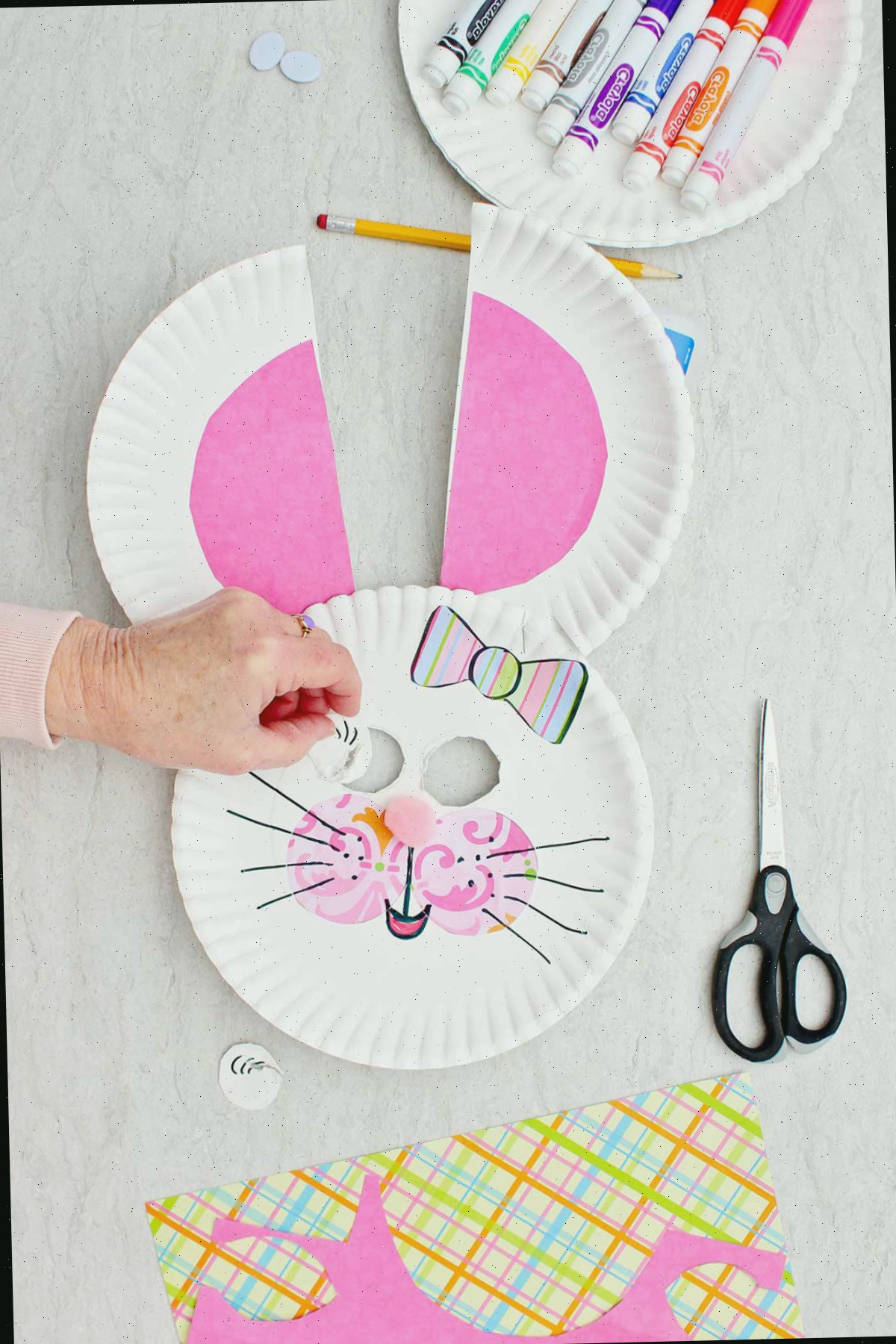 Indivíduo cortando e removendo aberturas para os olhos de um coelho de prato de papel para uma máscara, com uma placa de marcadores e outros materiais nas proximidades.