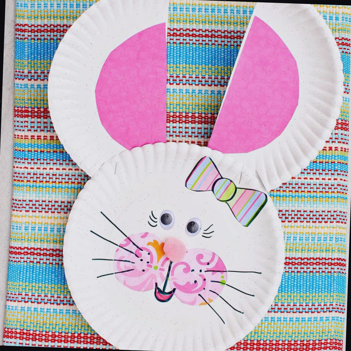 Coelhinho de prato de papel com orelhas e bochechas rosas, nariz de pom pom, laço listrado e olhos googly.