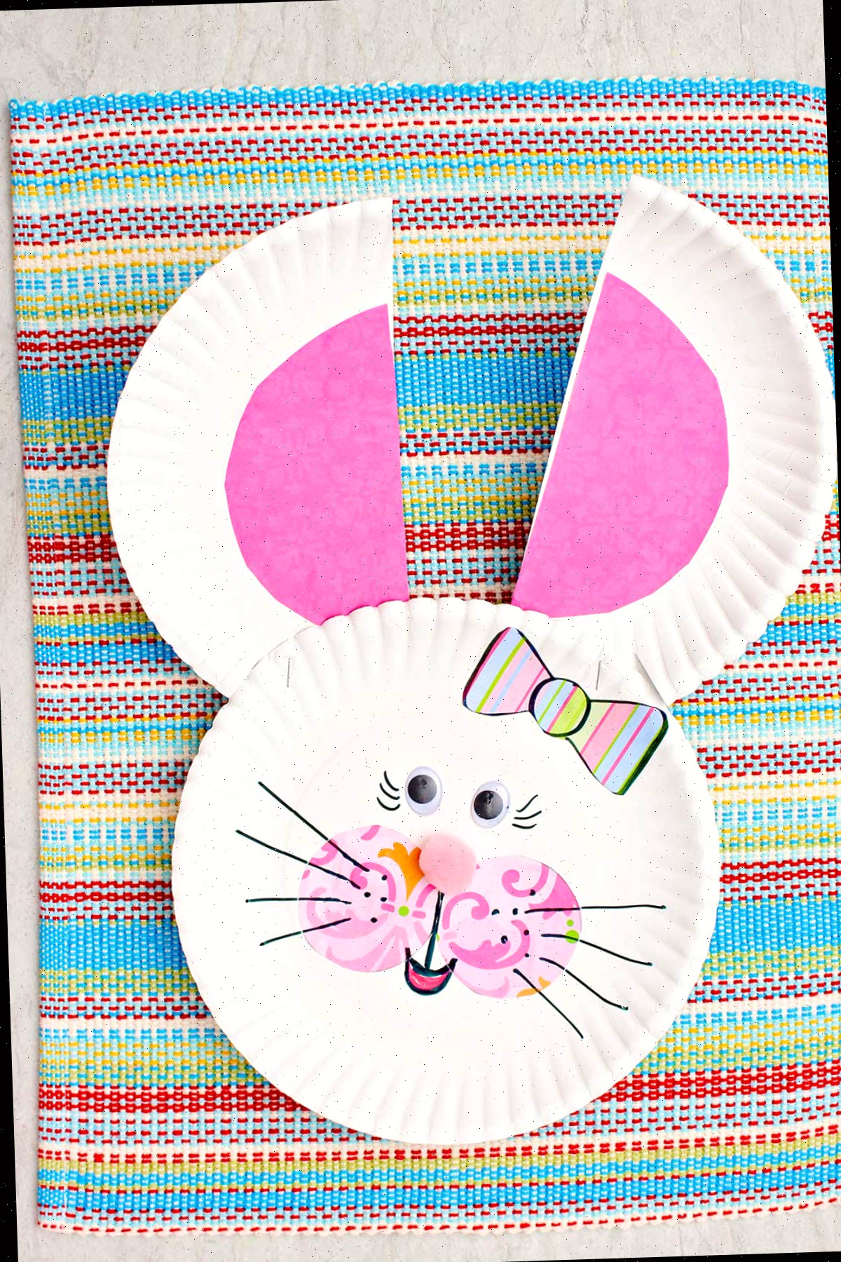 Coelhinho de prato de papel com orelhas e bochechas cor-de-rosa, nariz de pompom, laço listrado e olhos googly.
