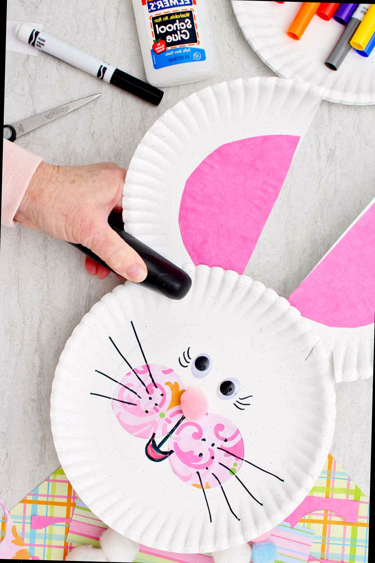 Indivíduo grampeando as orelhas em um prato de papel de coelho, que foram feitas cortando um prato de papel ao meio e anexando uma meia-lua rosa interna em cada uma.