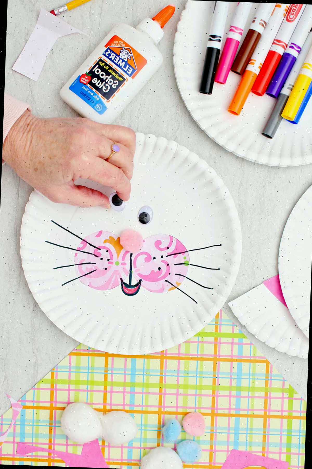 Indivíduo colocando um olho googly em um grande prato de papel para criar o rosto de um coelho, com outros materiais ao lado.