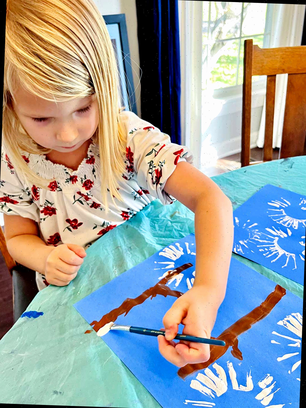 Menina loira com camisa floral adiciona tinta branca simulando neve em galhos em seu adorável artesanato de impressão de mão de cardeal de inverno.