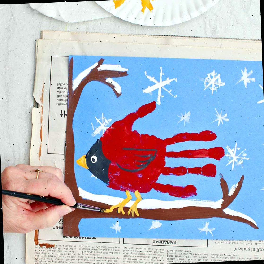 Pessoa pintando os detalhes finais em uma pintura de impressão de mão de cardeal inverno fofa de uma impressão de mão de cardeal sentada em um galho coberto de neve.