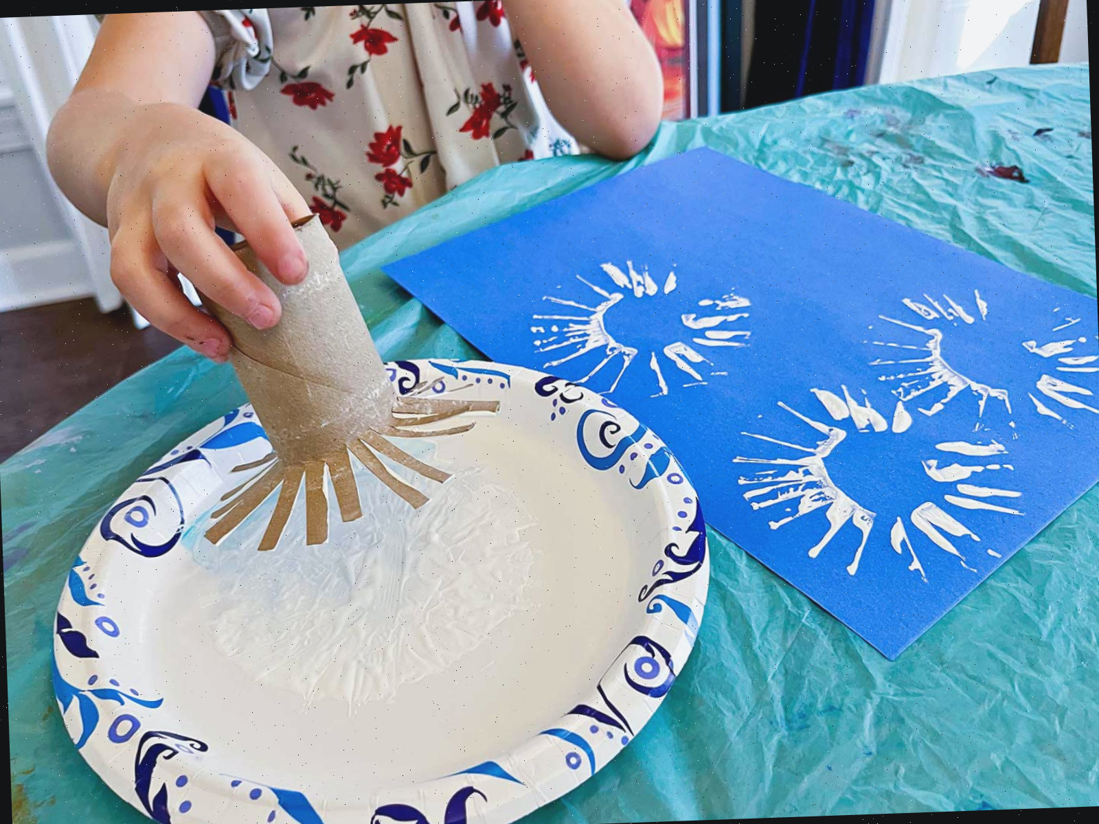Menina pequena usando um rolo de toalha de papel cortado como um sol para fazer carimbos para seu projeto em uma folha de papel azul.