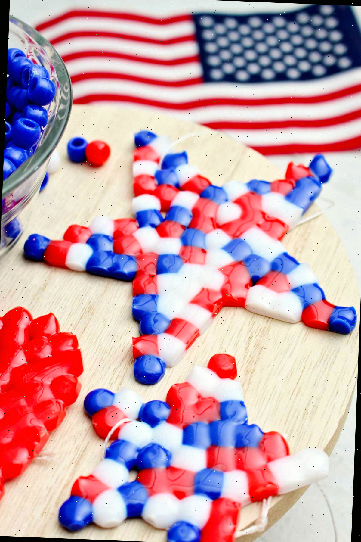 Duas pérolas derretidas finalizadas, nas cores vermelha, branca e azul, em forma de estrela, dispõem-se sobre um círculo de madeira ao lado de uma tigela com pérolas azuis e uma pequena bandeira americana ao fundo.