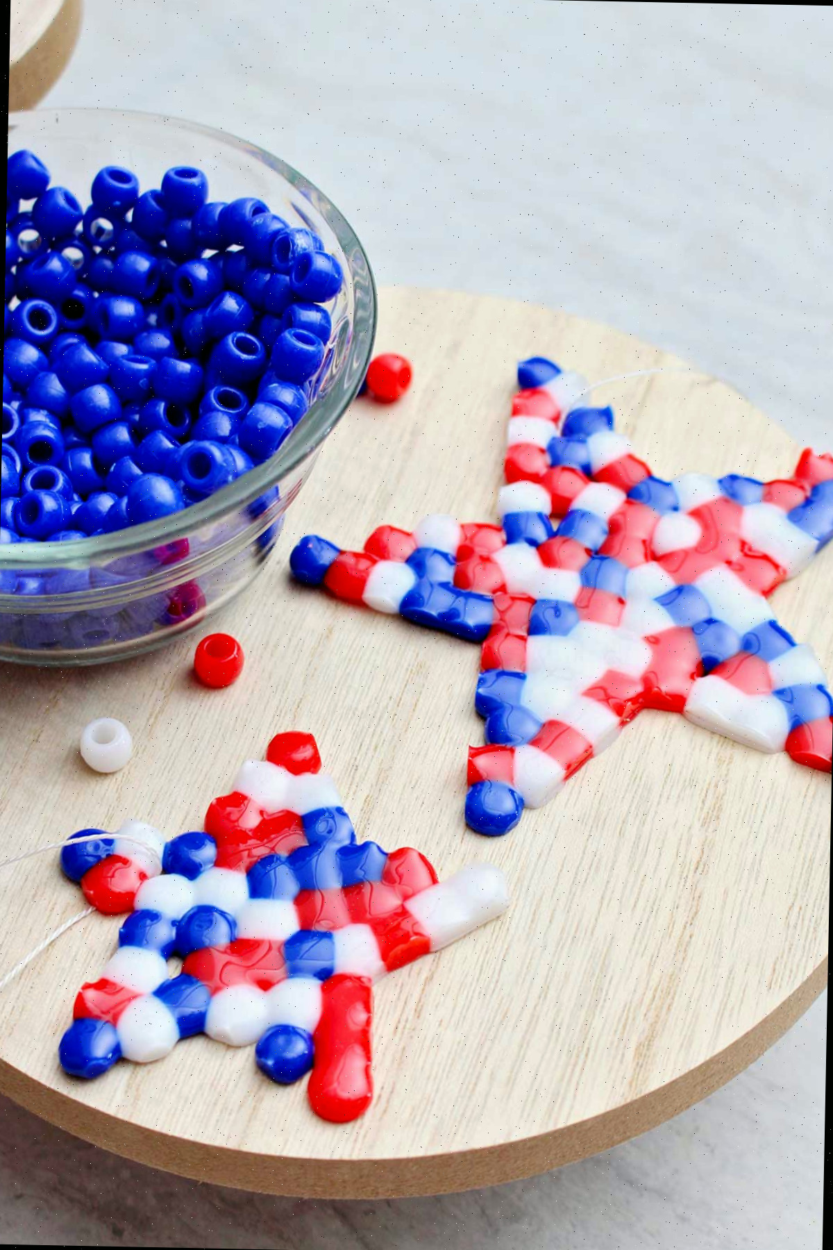 Duas contas derretidas completas em vermelho, branco e azul com formato de estrela posicionadas sobre um pedaço circular de madeira ao lado de uma tigela de contas azuis.