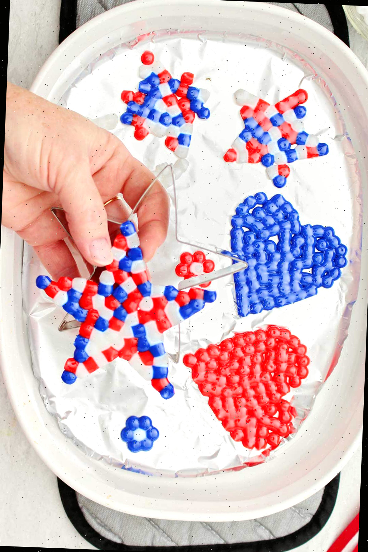 Uma pessoa retirando uma estrela de contas derretidas nas cores vermelha, branca e azul de um cortador de biscoitos em formato de estrela, com outros projetos de contas derretidas ao fundo.