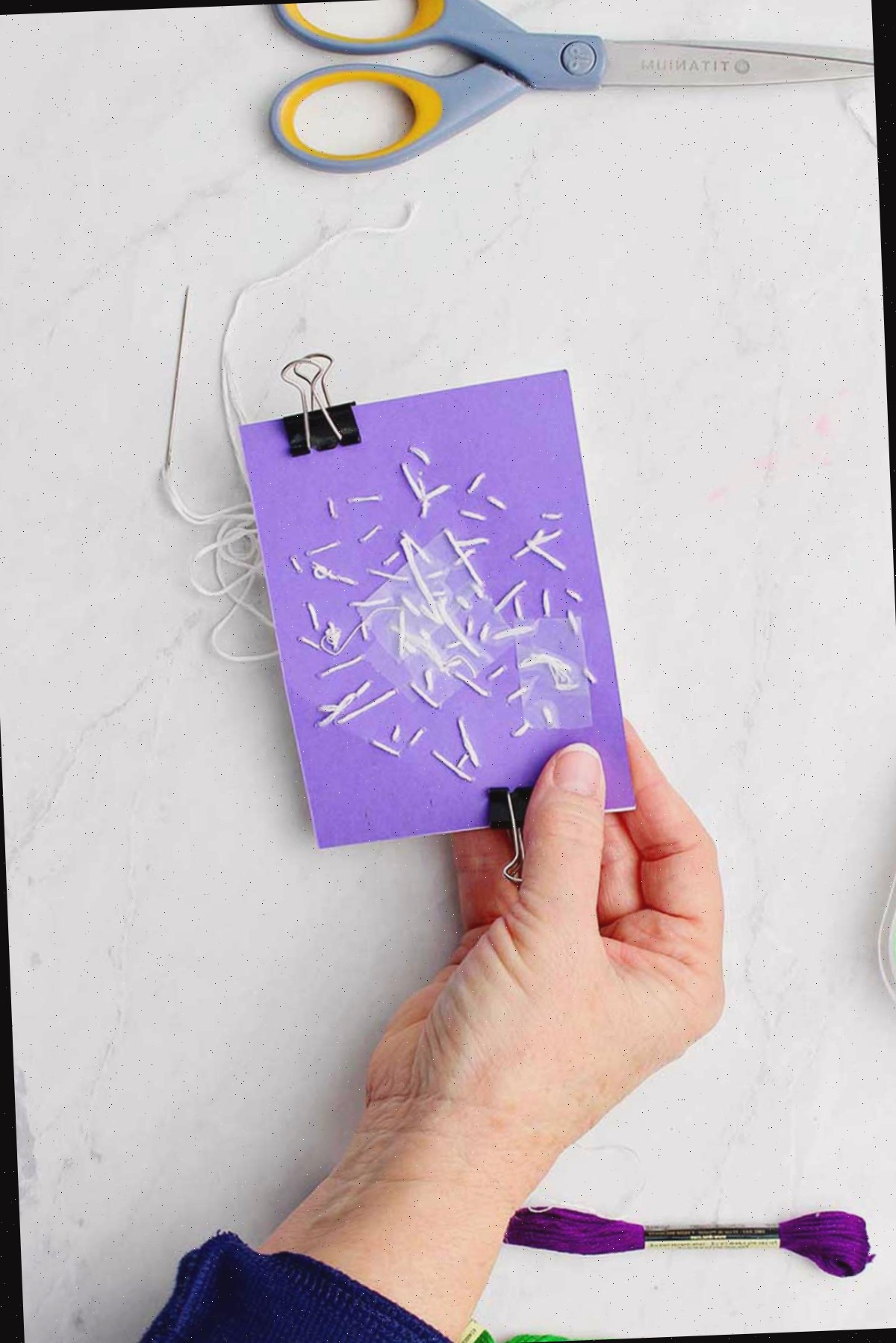 Mão segurando atrás do papel roxo mostrando os pontos de bordado.