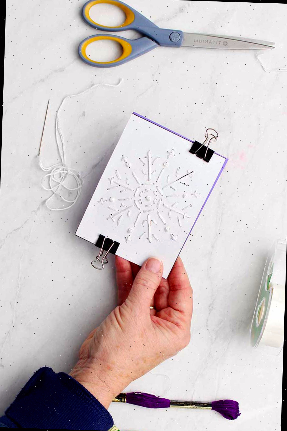 Mão segurando papel de floco de neve bordado preso em cartão roxo com fio de bordado, fita e tesoura próximas.