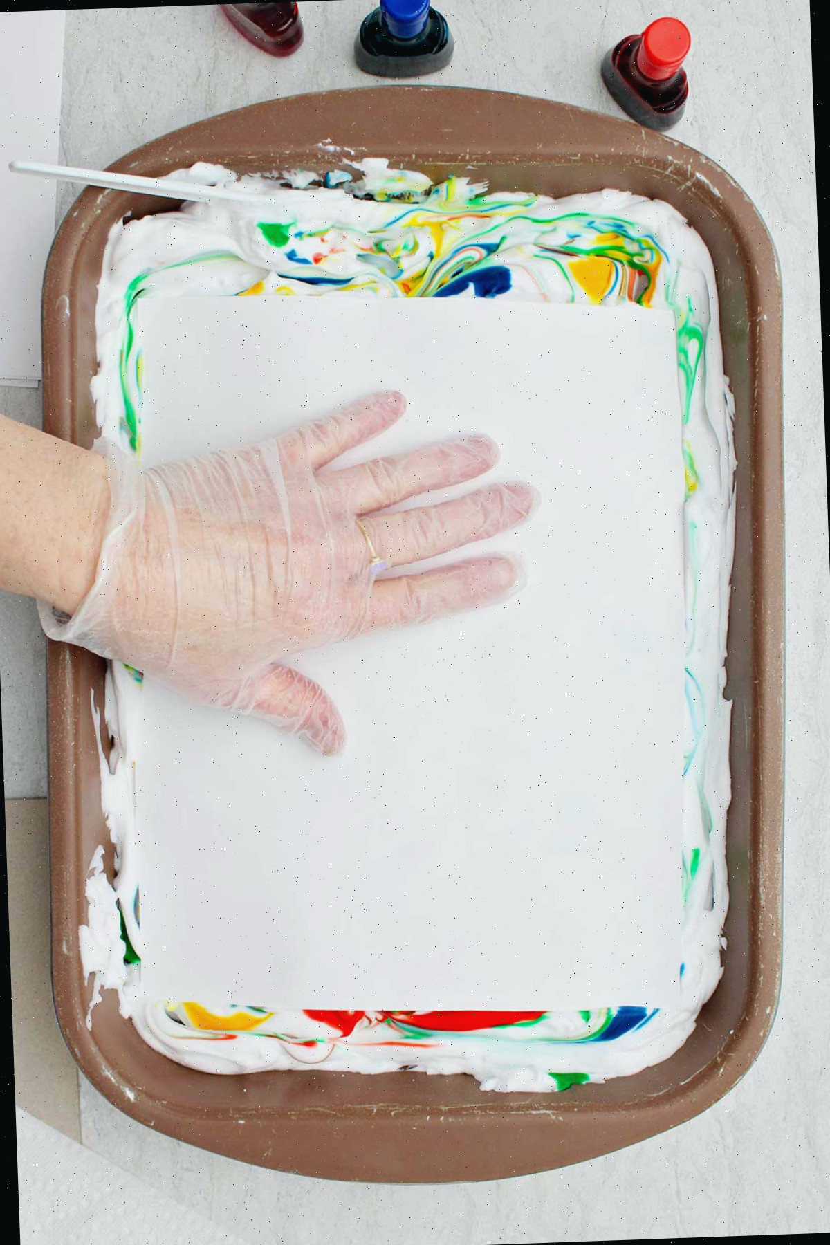 Indivíduo utilizando luvas plásticas pressionando um pedaço de papel contra o creme de barbear colorido.