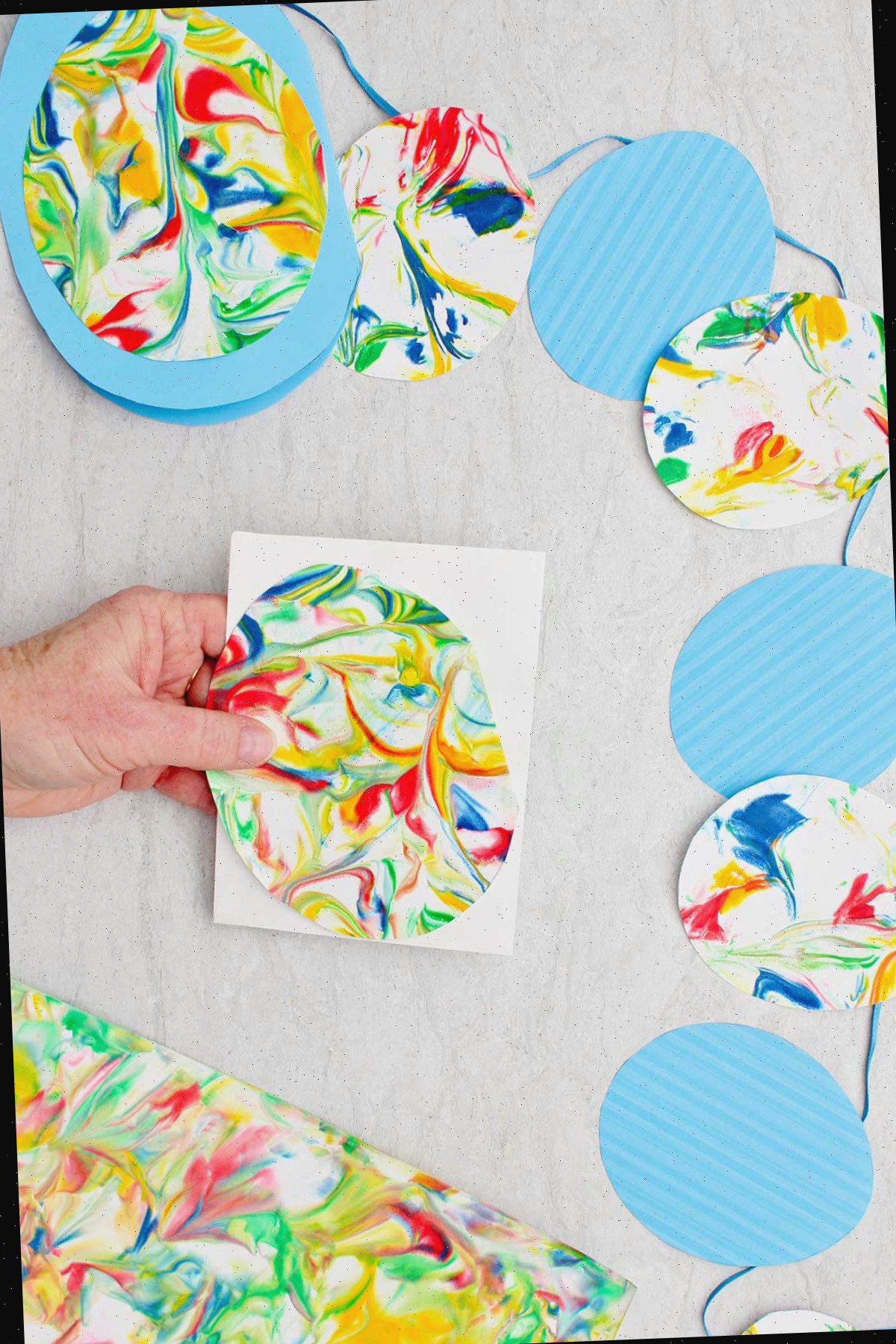 Pessoa segurando um ovo marmorizado em um pedaço de papel branco com bandeirinhas de ovos de Páscoa feitos de ovos marmorizados e ovos de papel azul por perto.