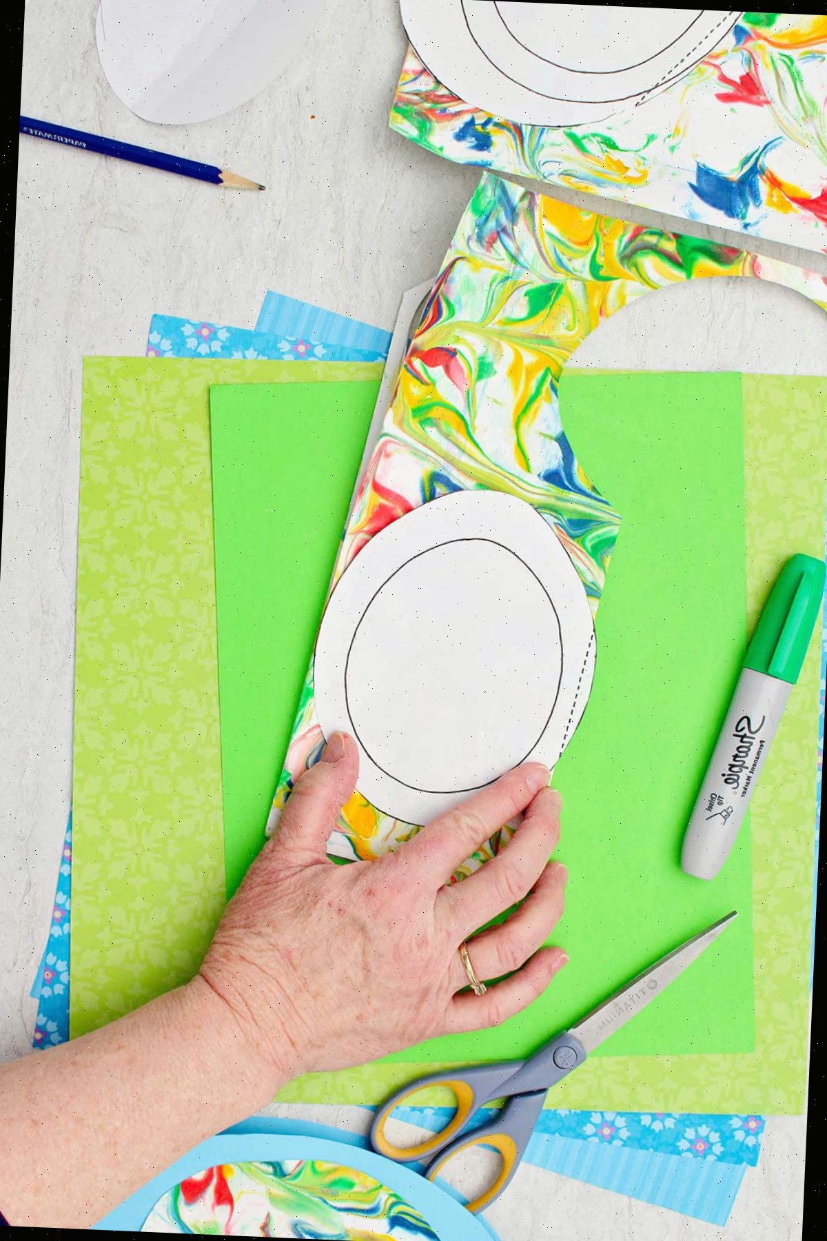 Indivíduo traçando um molde de ovo em papel colorido mármore com outros materiais próximos.