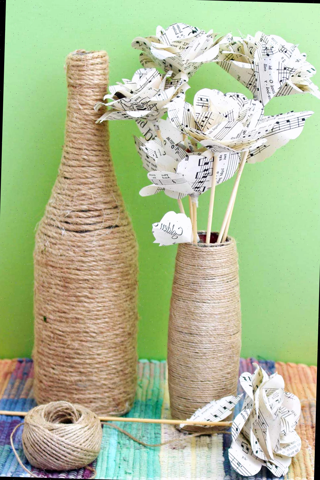 Uma garrafa coberta em fio e um vaso adornado com fio contendo flores de papel, sobre uma mesa colorida contra um fundo verde.