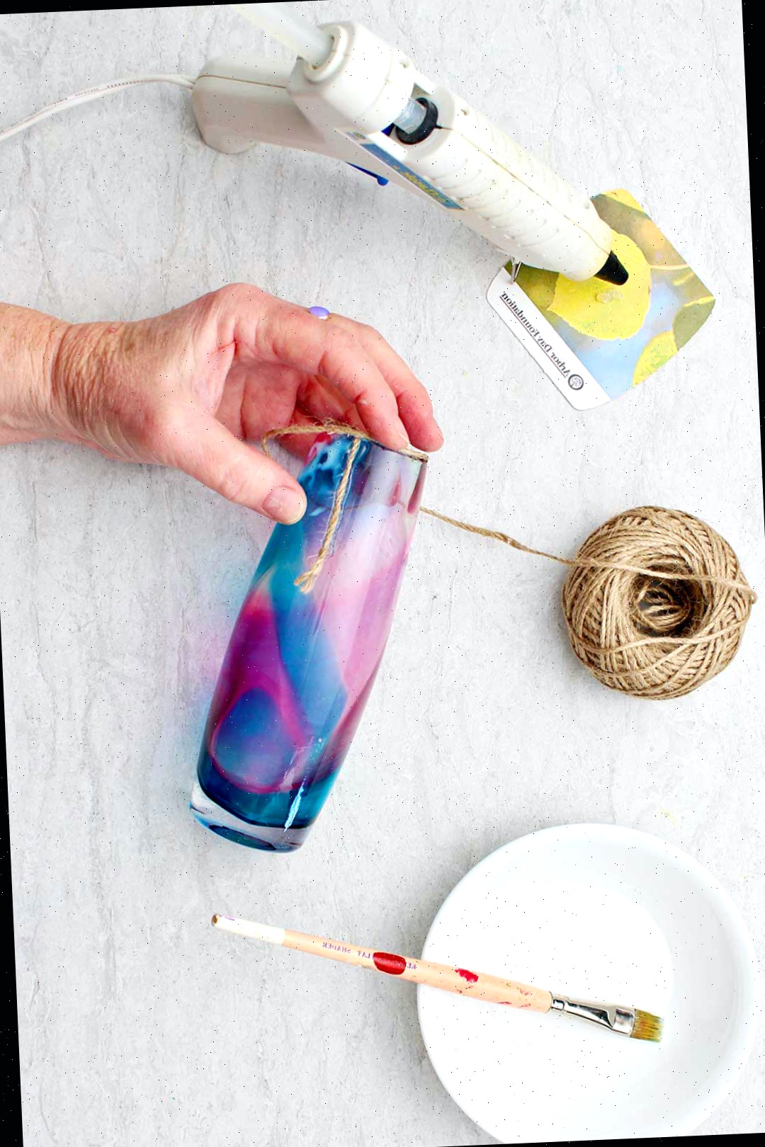 Mão segurando vaso de vidro com o início do envoltório de fio colado a ele com pistola de cola e cola branca, com pincel próximo.