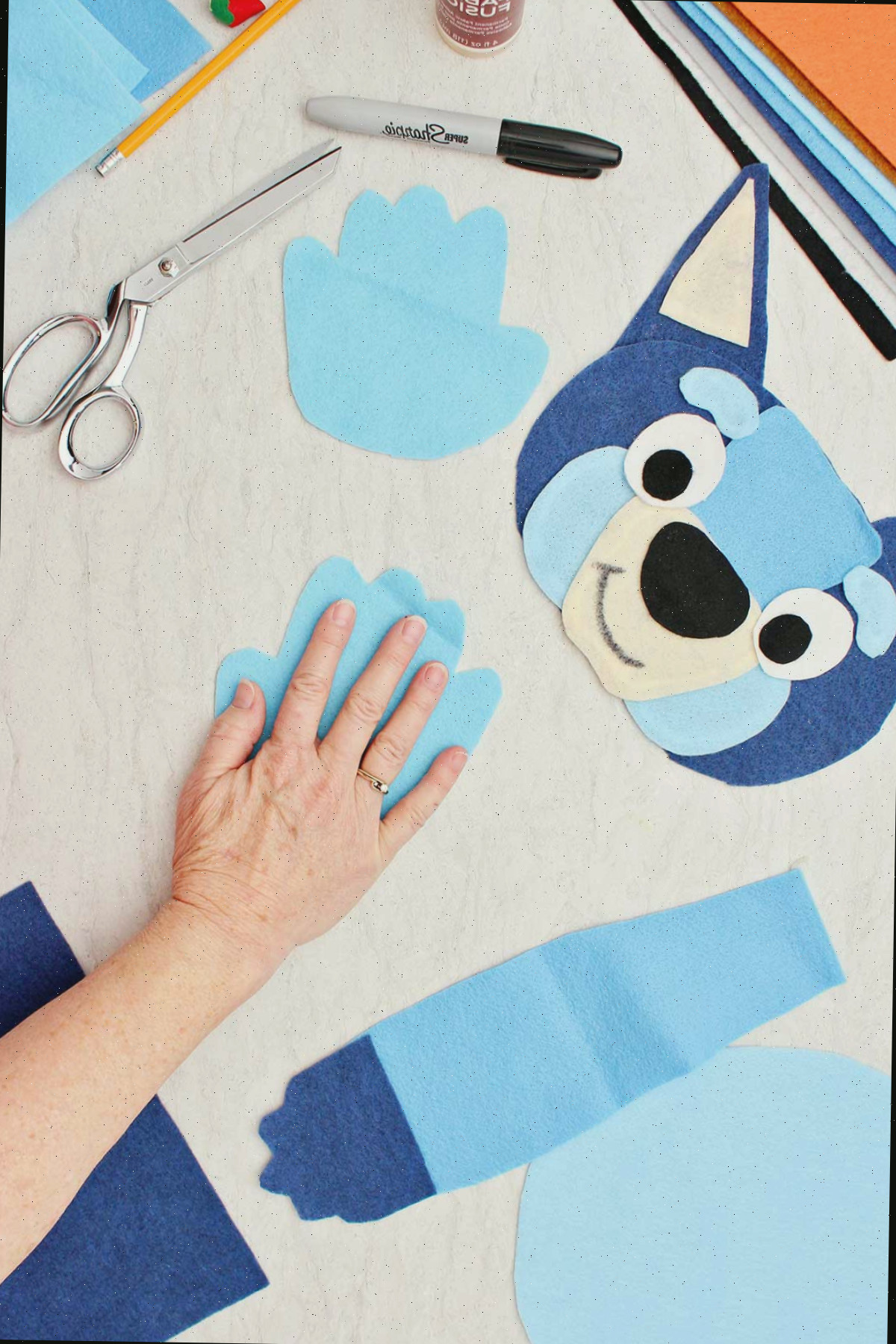 Pessoa medindo sua mão para criar uma pata azul para a fantasia do Bluey com outras partes ao redor.