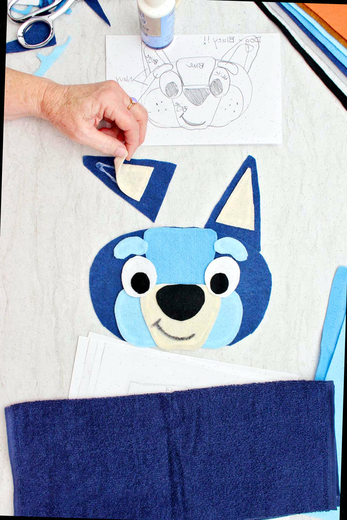 Colando à mão a parte interna branca da orelha para a fantasia do Bluey com outras peças da fantasia ao redor.