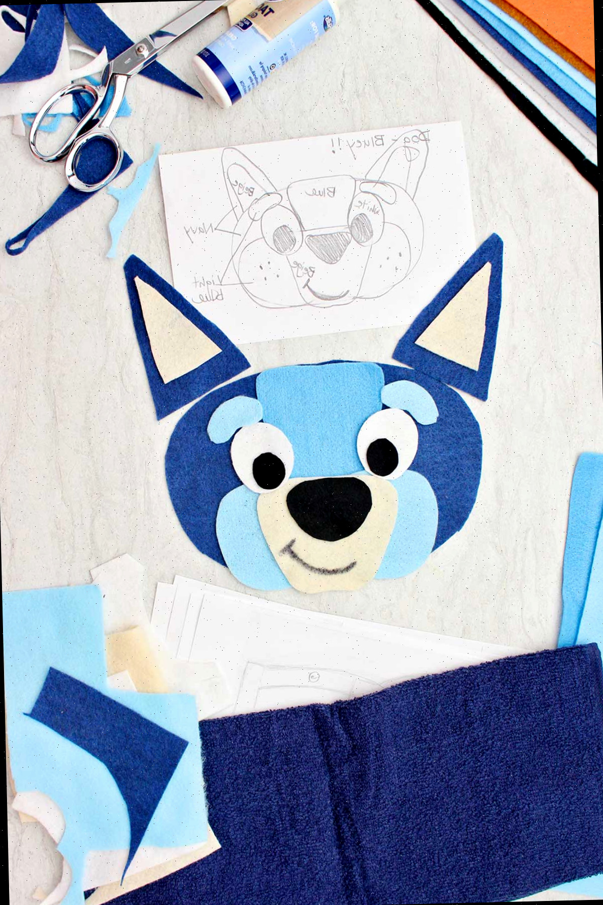 Um esboço para uma fantasia do Bluey próximo a alguns pedaços cortados de feltro para o rosto e outros retalhos de materiais ao redor.