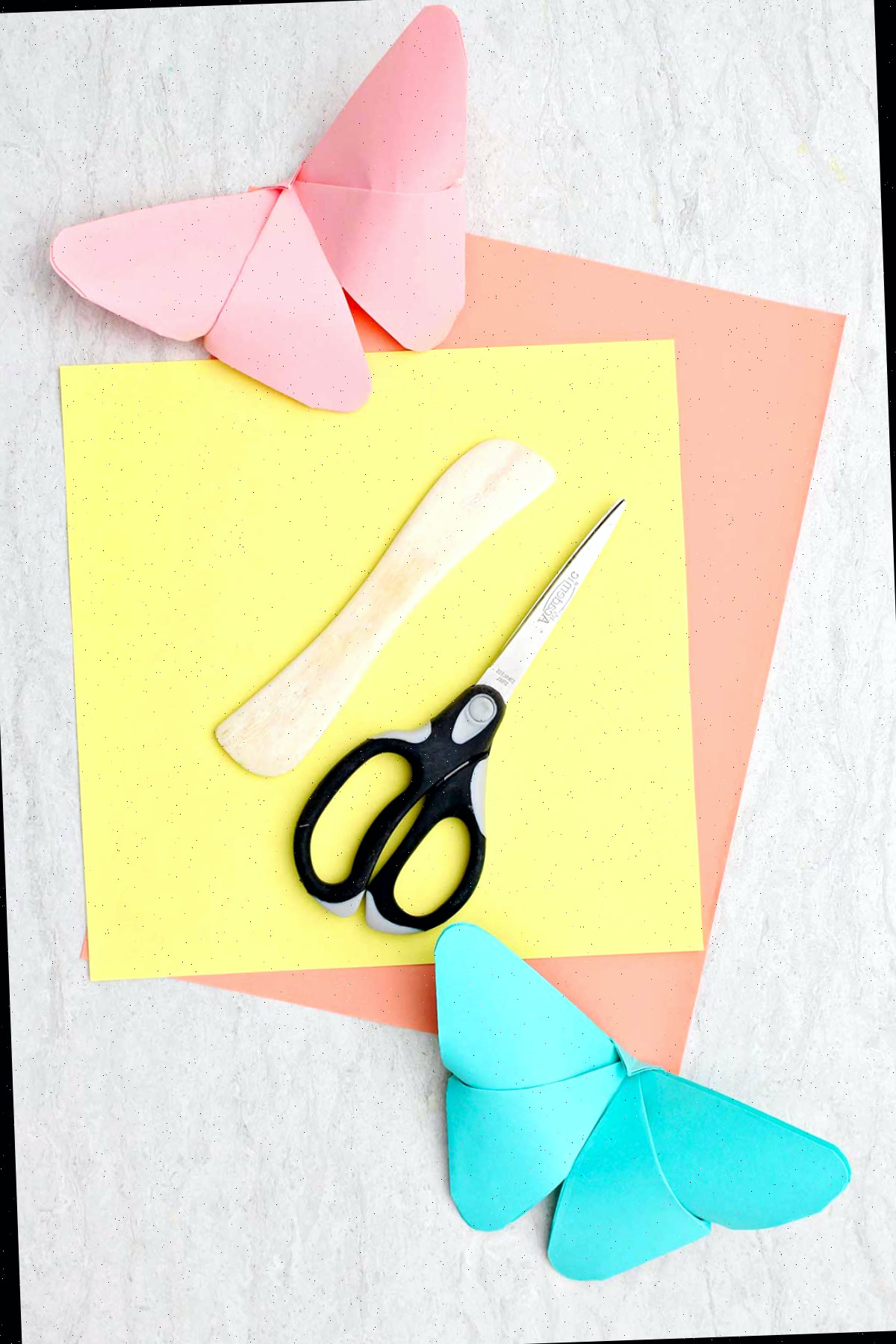 Uma borboleta de origami rosa e azul claro, um pedaço de papel amarelo e laranja, um vincador de papel de madeira e uma tesoura.