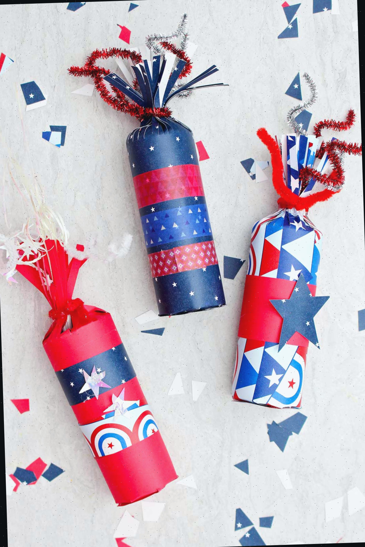 Três caixas de doces em forma de foguete ornamentadas com papel vermelho, branco e azul e fita washi, com alguns pedaços de papel festivo espalhados ao redor.
