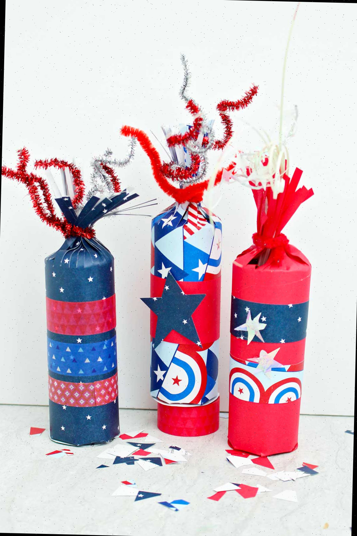 Vista direta de três caixas de guloseimas em forma de fogos de artifício decoradas com papéis vermelhos, brancos e azuis e fita washi, com alguns retalhos de papel festivo espalhados ao redor.