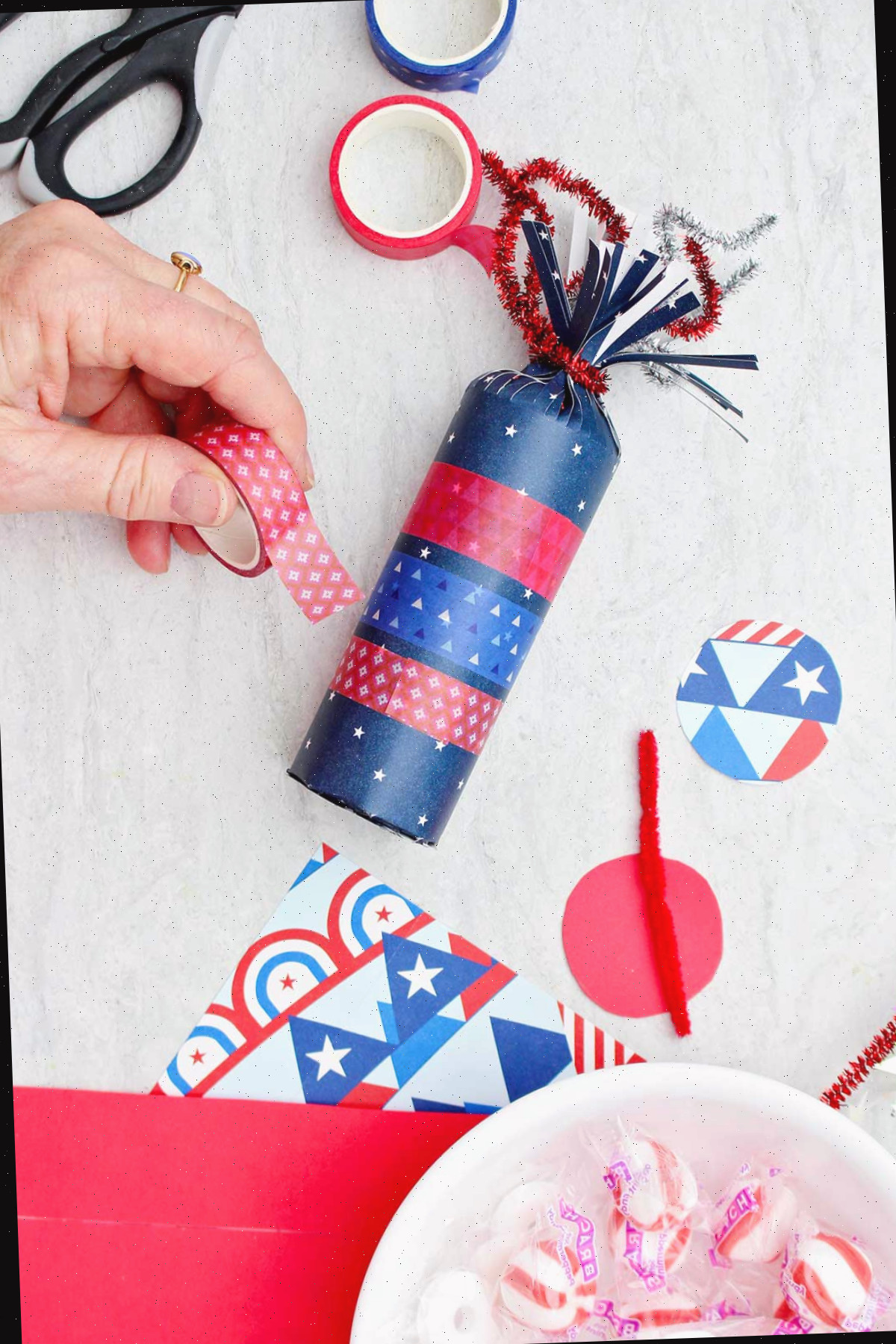 Indivíduo decorando a caixa de doces em forma de foguete azul com fita washi vermelha.