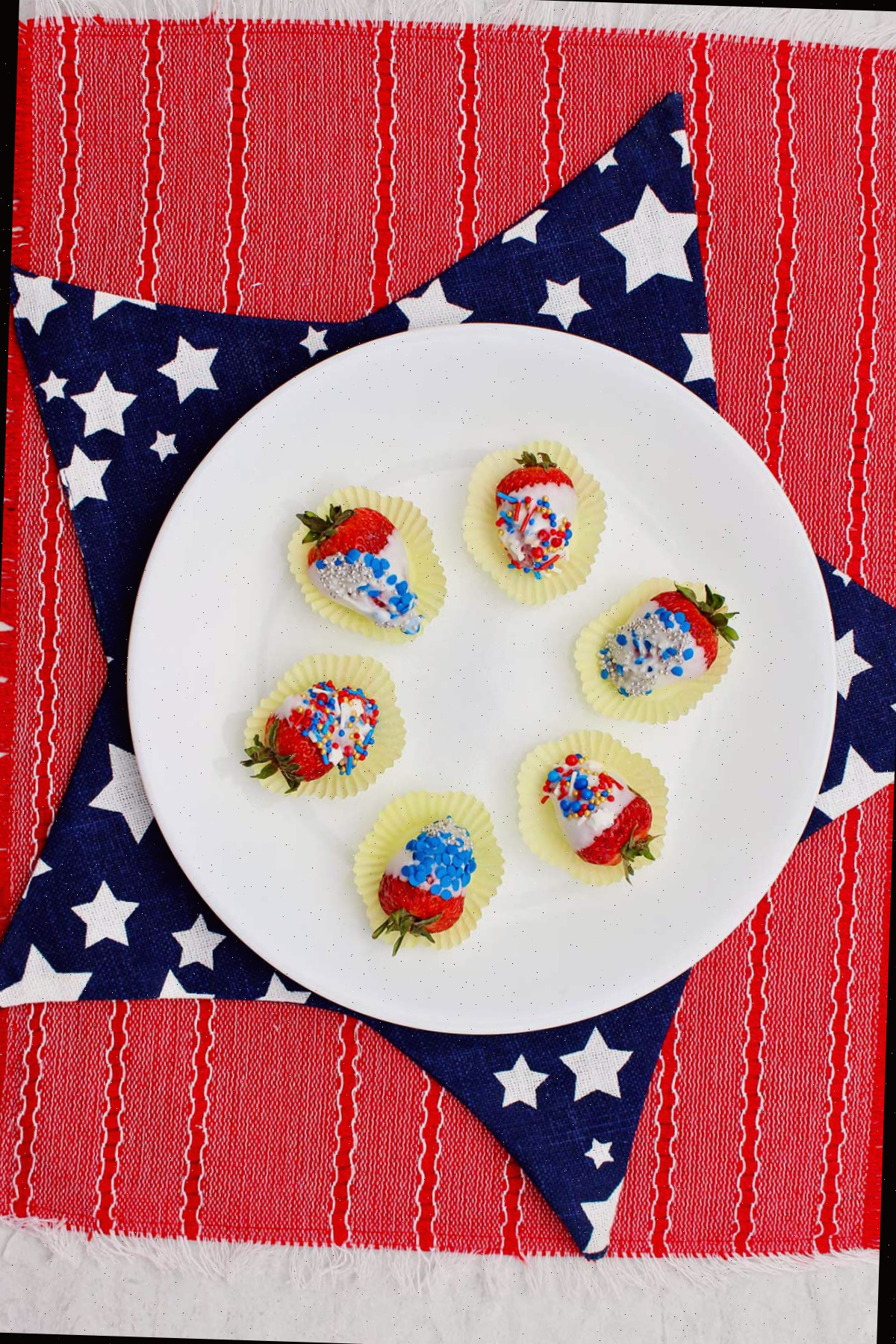 Prato branco com morangos submersos em chocolate branco finalizados sobre um pano de estrelas azul e branco e uma toalha de mesa vermelha.