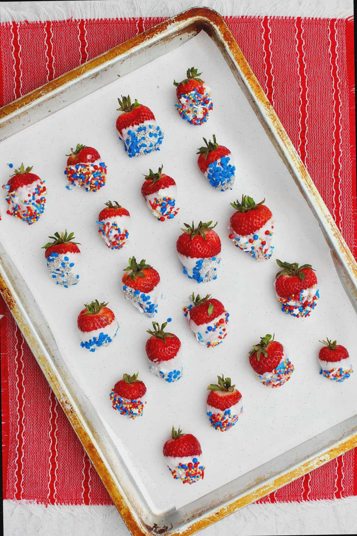 Morangos banhados em chocolate branco decorados com confetes azuis, vermelhos e prateados em uma assadeira com papel manteiga.