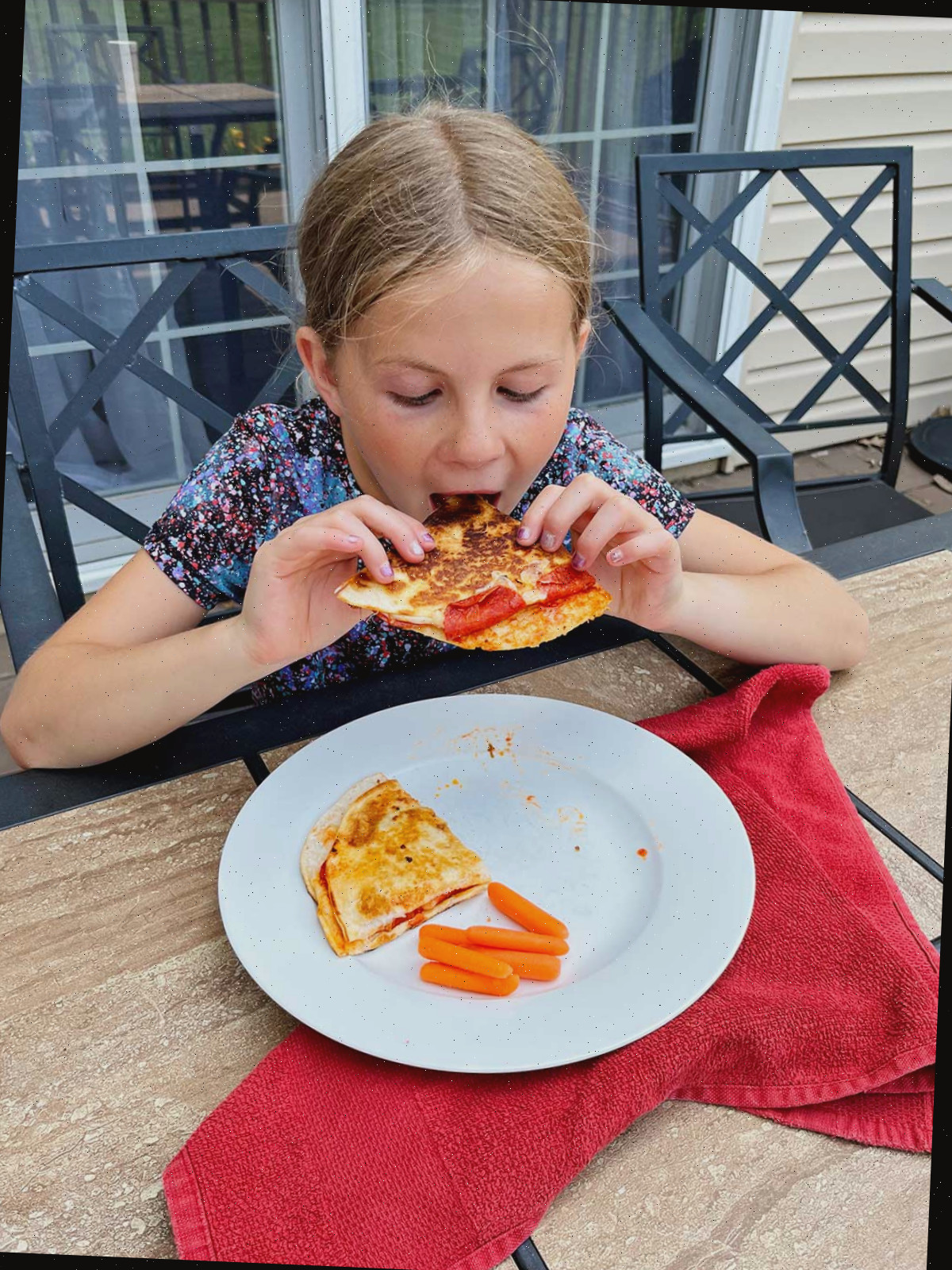Uma menina jovem com cabelo loiro comendo uma quesadilla de pizza em uma mesa ao ar livre.
