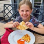 Uma menina jovem com cabelo loiro sorrindo para a câmera e segurando uma quesadilla de pizza em uma mesa ao ar livre.