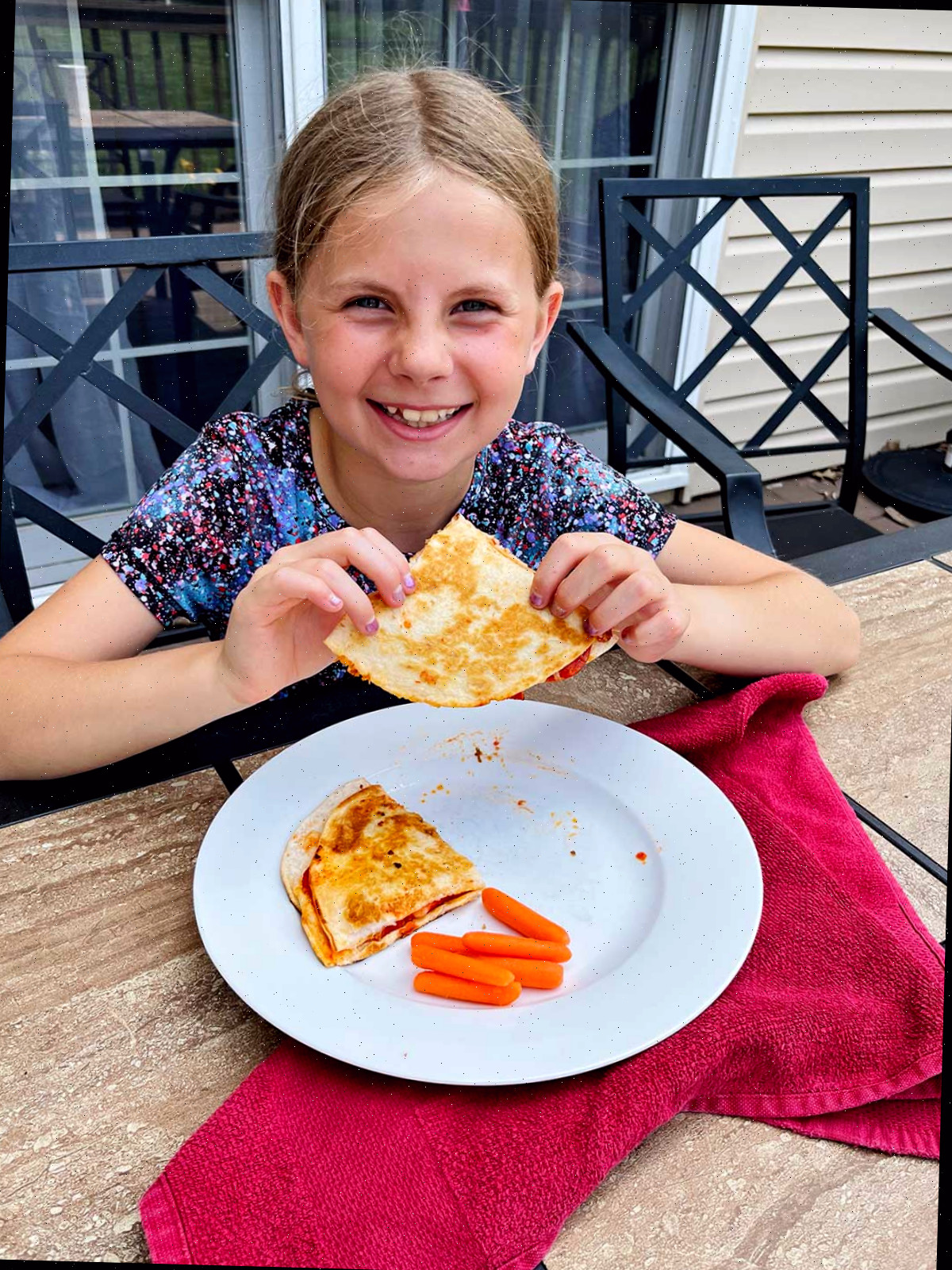Uma menina com cabelo loiro sorrindo para a câmera e segurando uma quesadilla de pizza em uma mesa ao ar livre.