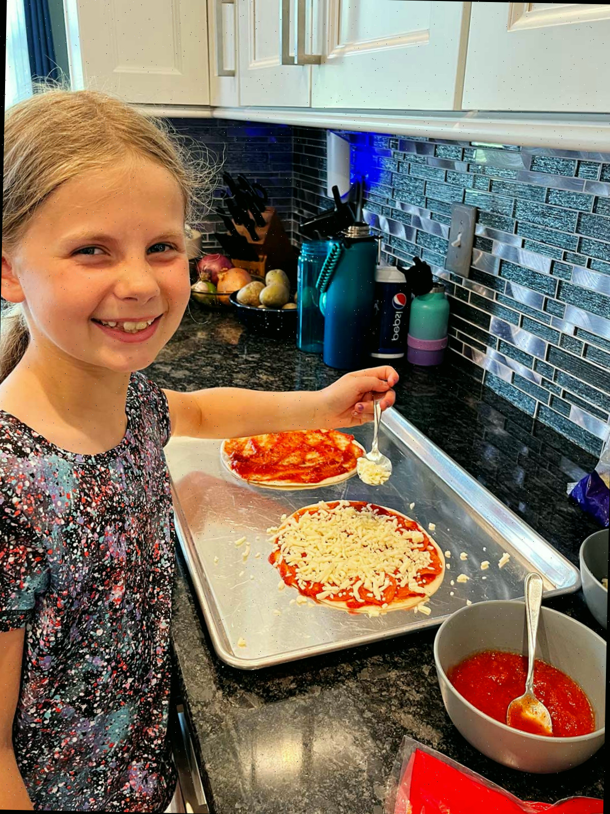 Uma menina jovem, sorrindo para a câmera, montando duas quesadillas de pizza na bancada.