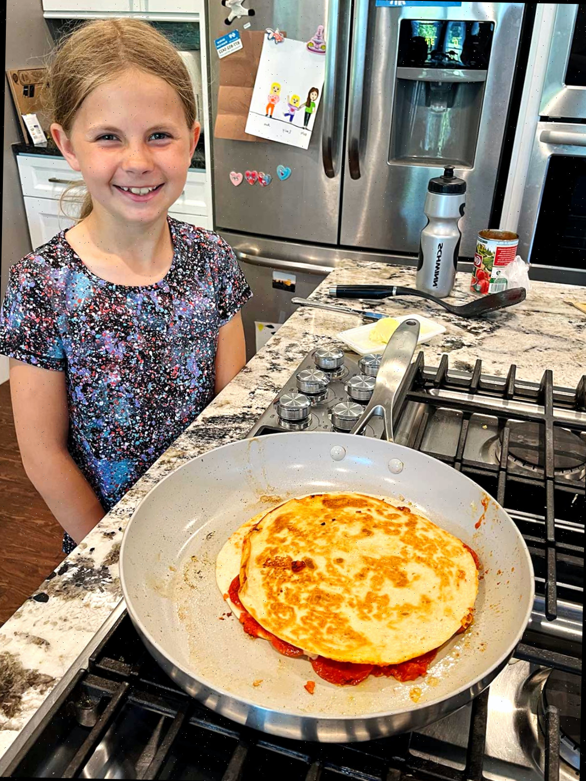 Uma menina jovem com cabelo loiro sorri para a câmera enquanto cozinha uma quesadilla de pizza no fogão.