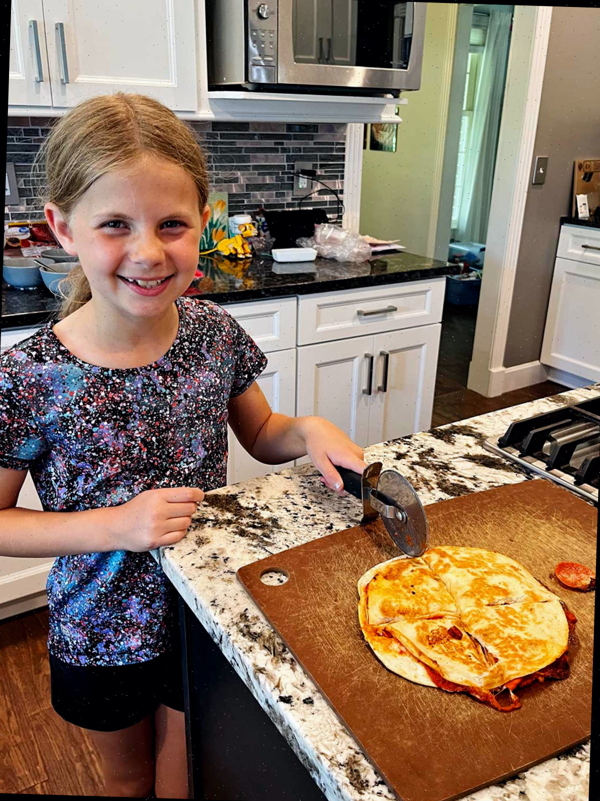 Uma menina jovem com cabelo loiro sorrindo para a câmera e cortando uma quesadilla de pizza em quatro partes na cozinha.