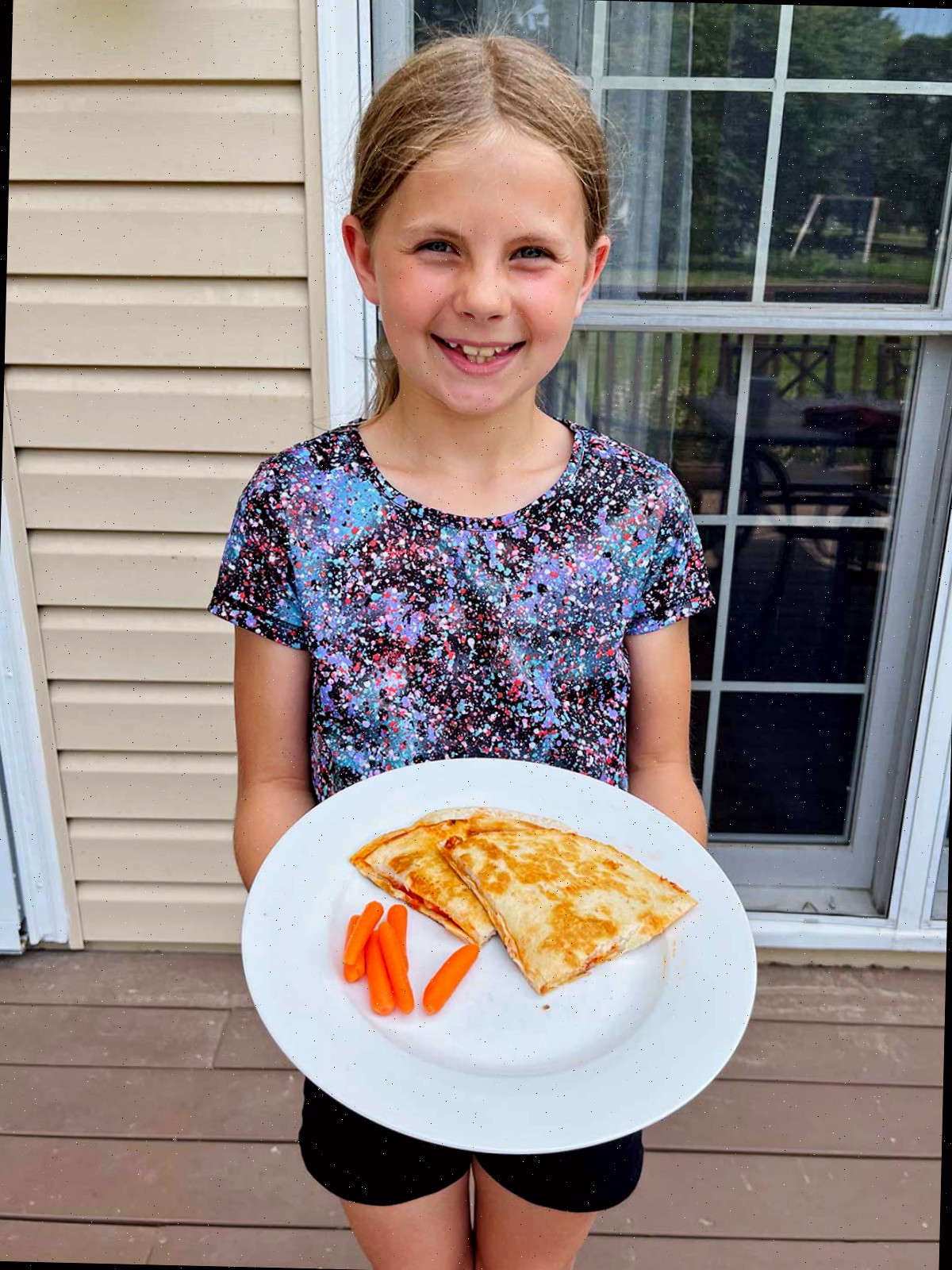 Uma jovem menina sorridente com cabelo loiro segurando um prato branco com duas quesadillas de pizza e cenouras baby ao lado.