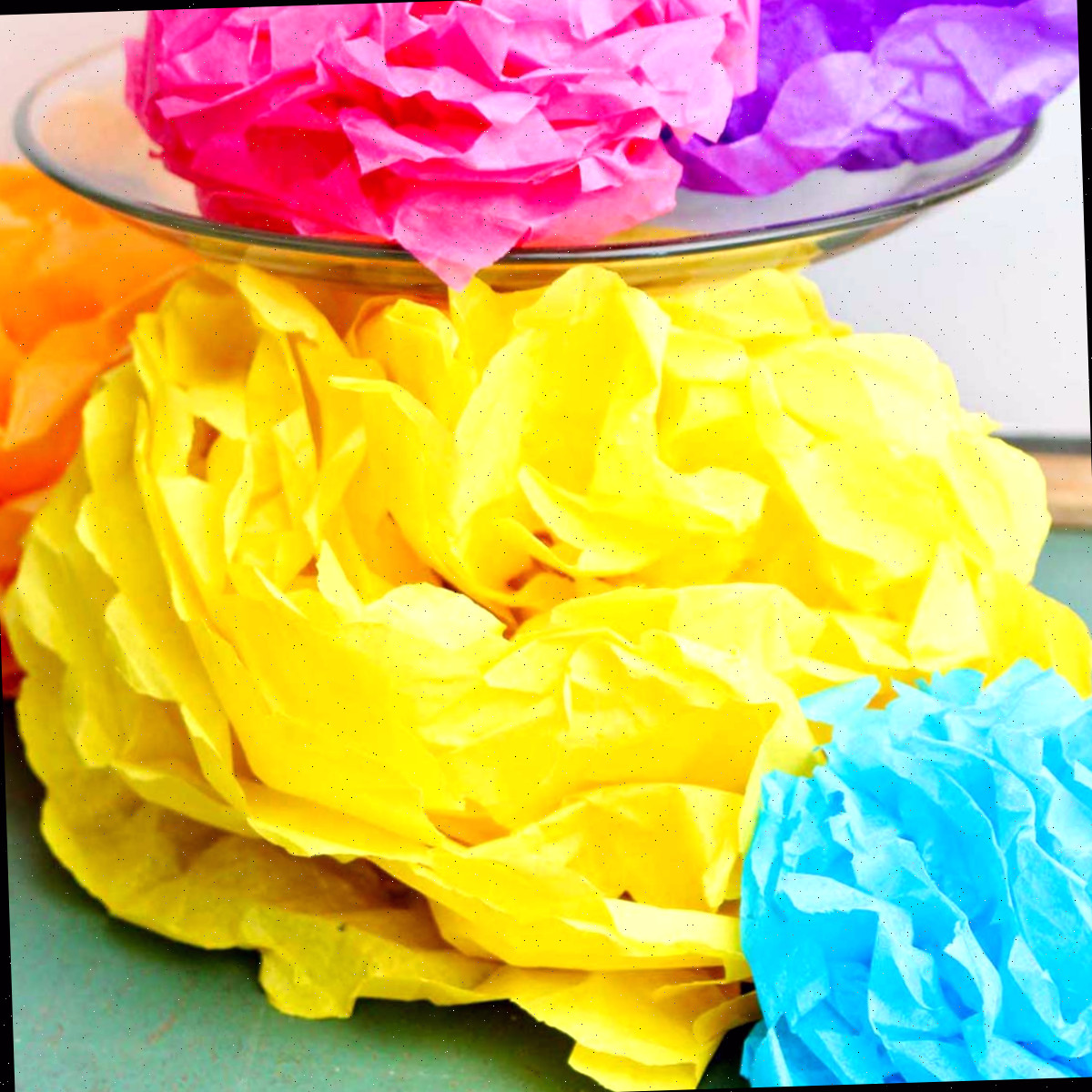 Flores de papel de seda coloridas concluídas, uma roxa e outra rosa descansando em um suporte de bolo de vidro com outras embaixo.