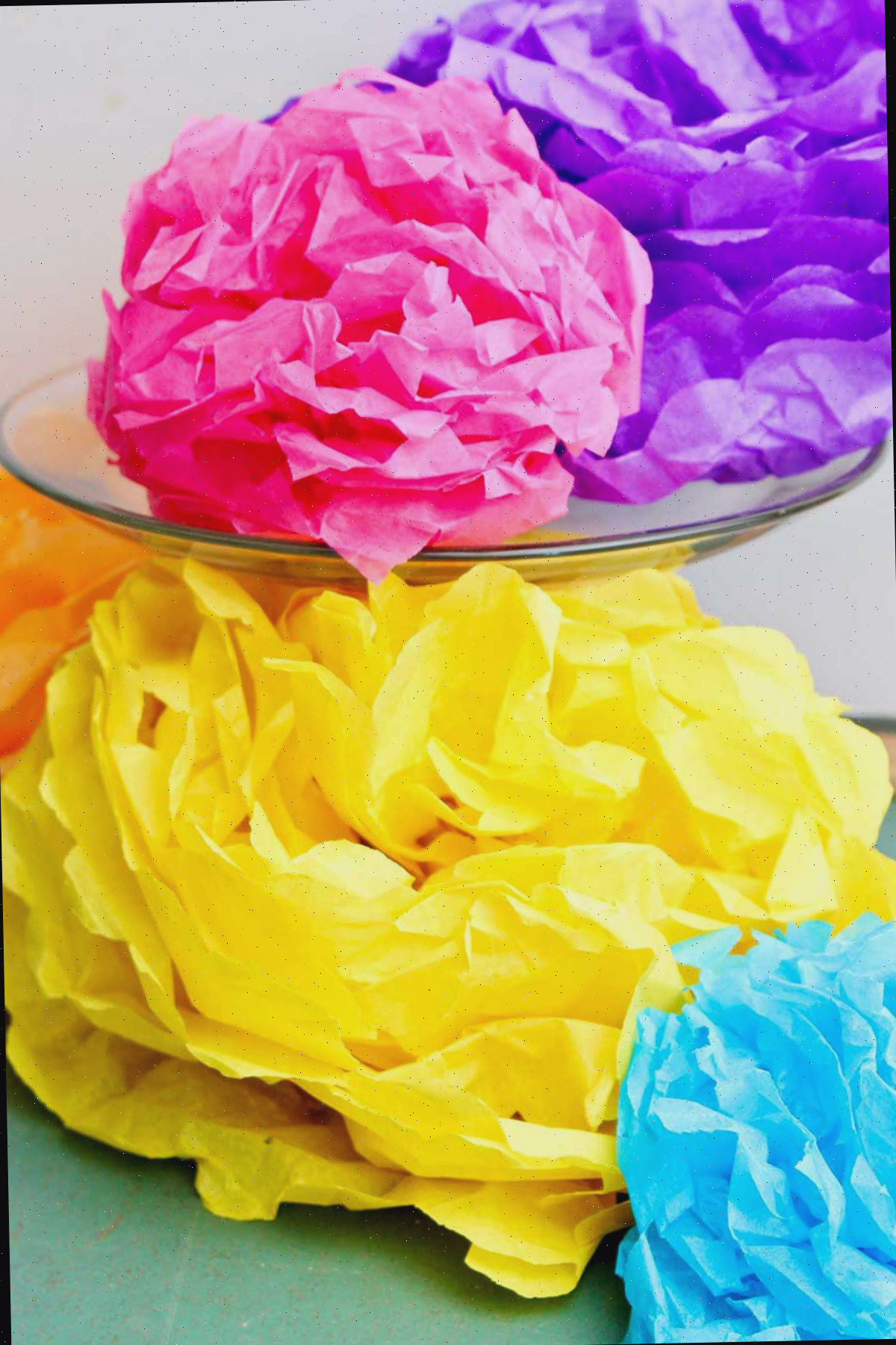 Flores de papel de seda coloridas finalizadas, uma roxa e rosa repousando em um display de vidro com outras abaixo.