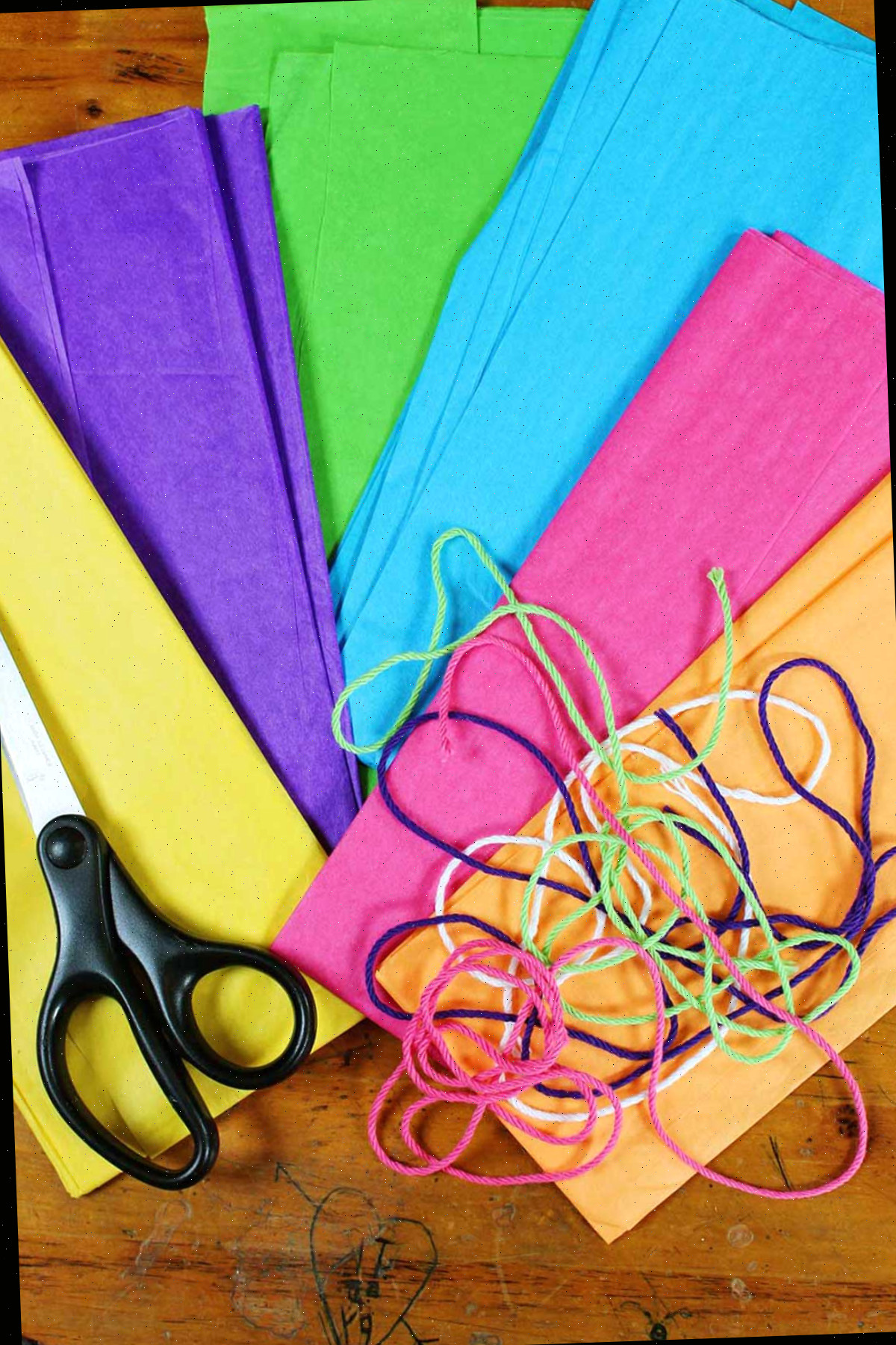 Pilhas de papel de seda laranja, rosa, azul, verde, roxo e amarelo com linha e tesoura descansando sobre eles.
