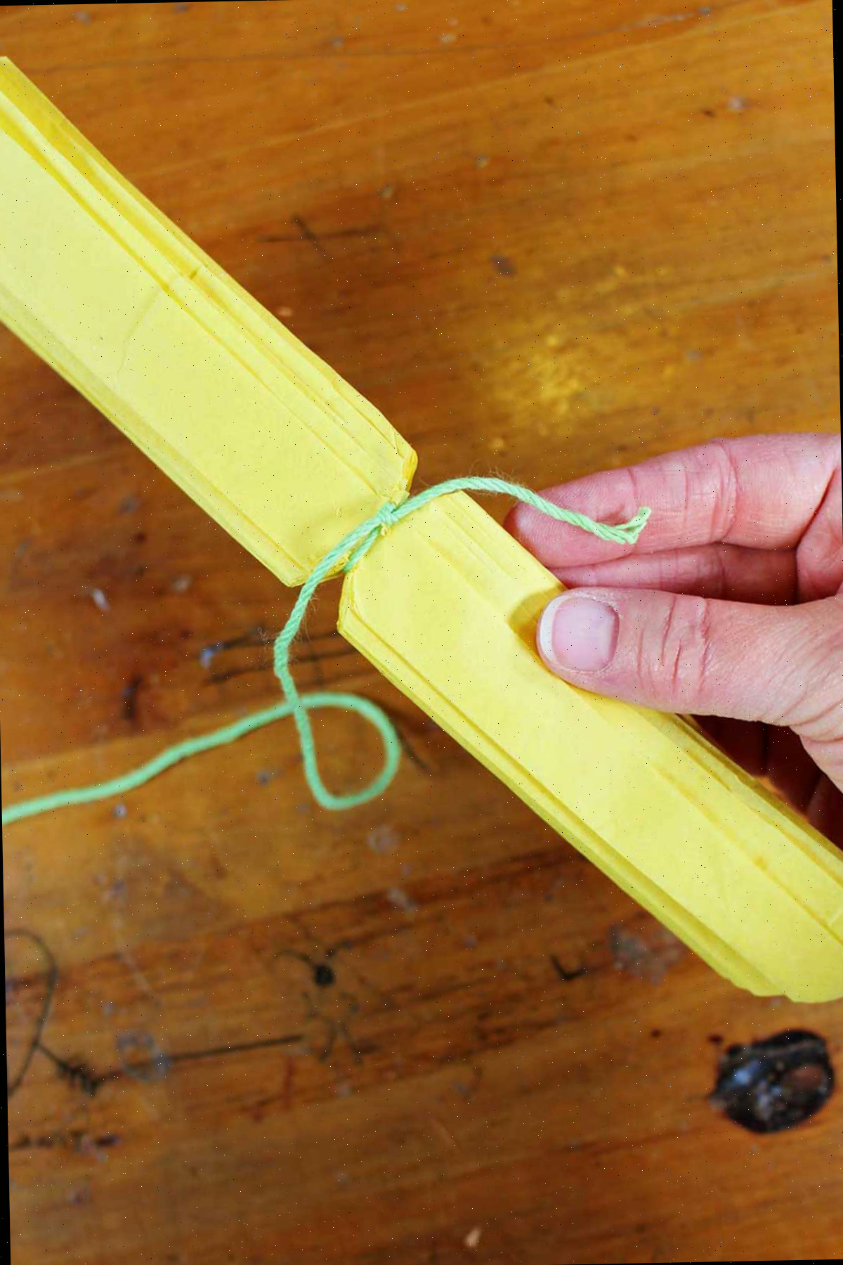 Mão segurando papel crepom amarelo dobrado e aparado com um fio verde amarrado ao redor do centro.