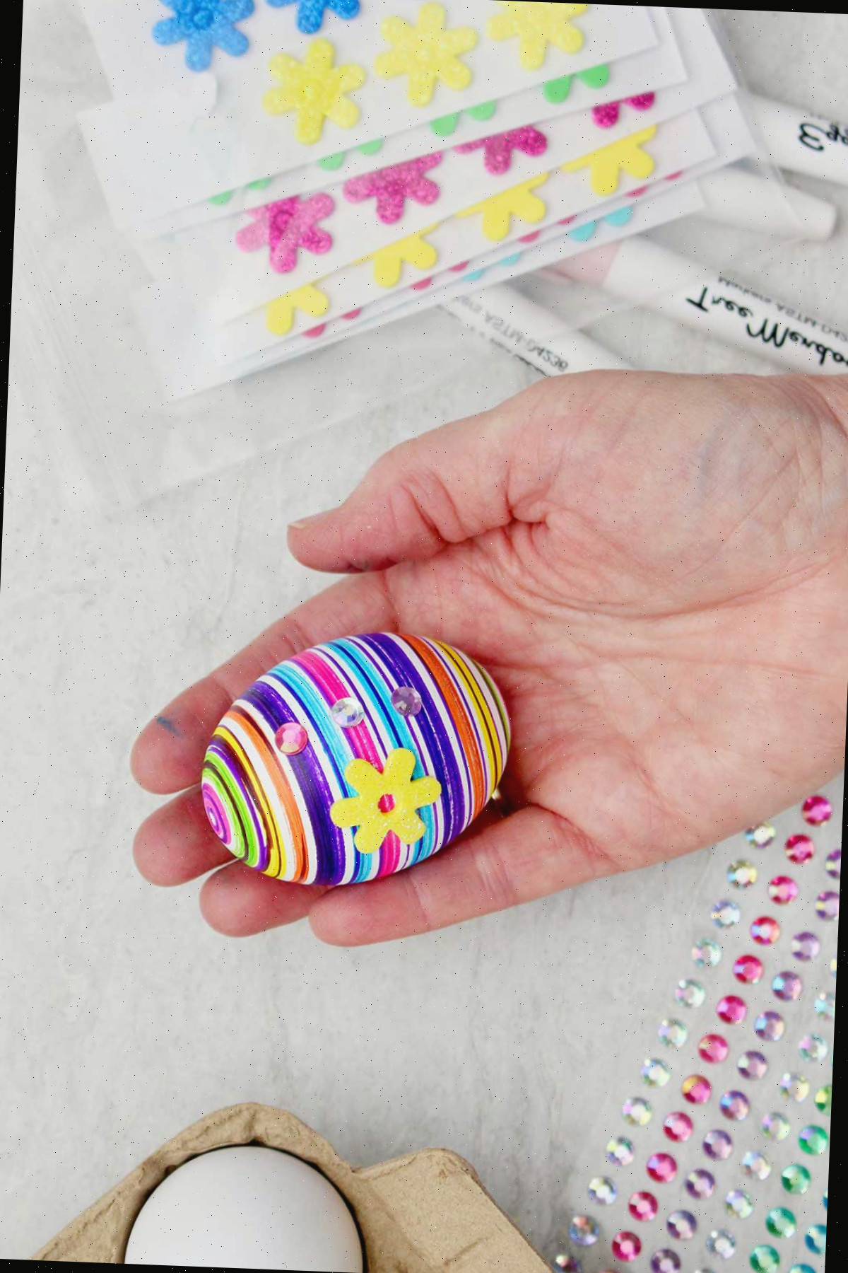 Uma pessoa segurando um ovo totalmente decorado com caneta, strass e uma flor amarela.