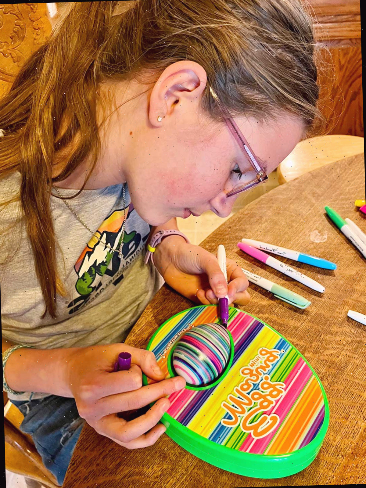 Uma menina em idade escolar com cabelo castanho claro pintando um ovo com uma máquina EggMazing em uma mesa de cozinha.